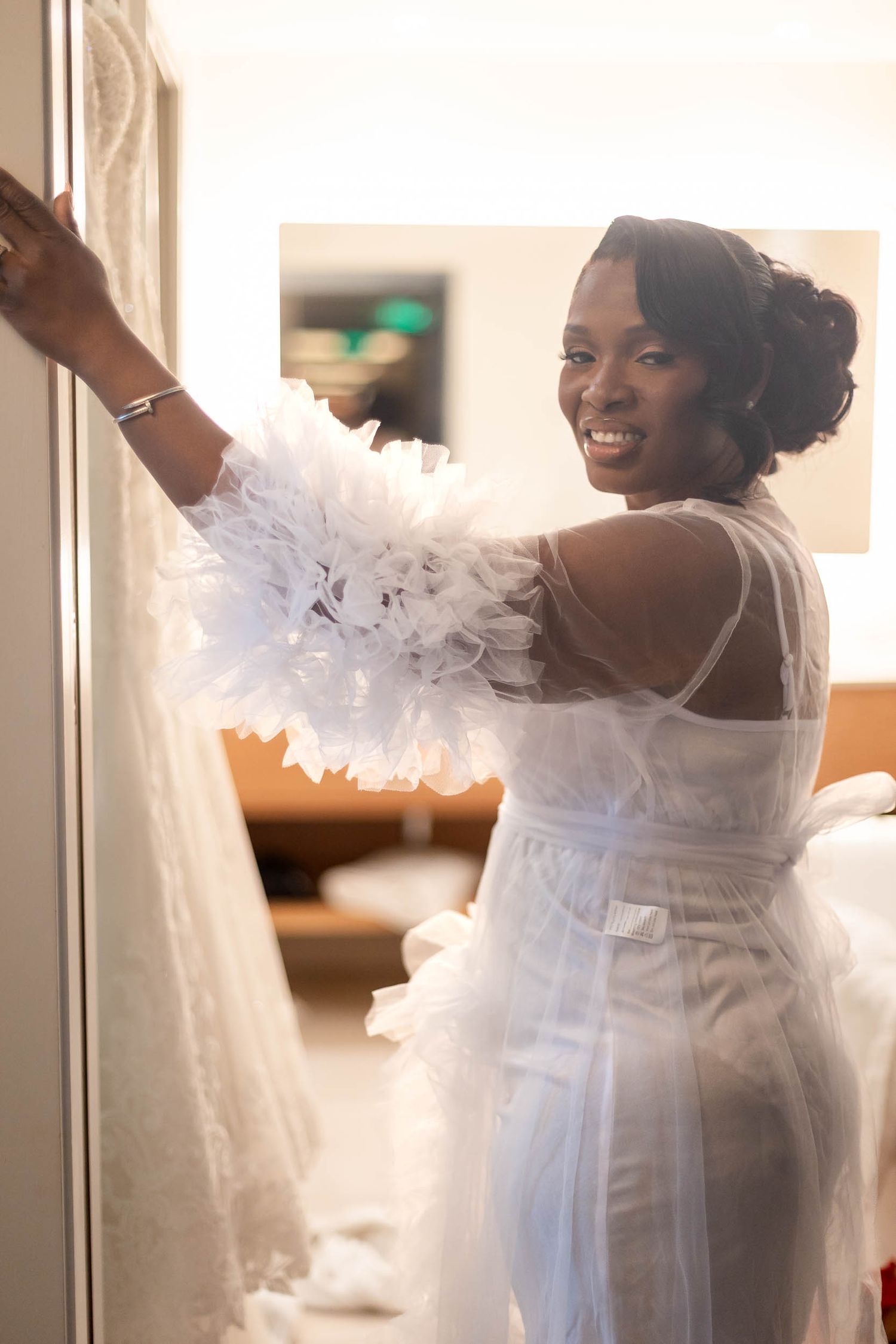Smiling bride in white feather boa and sheer gown holding her wedding dress before the ceremony.