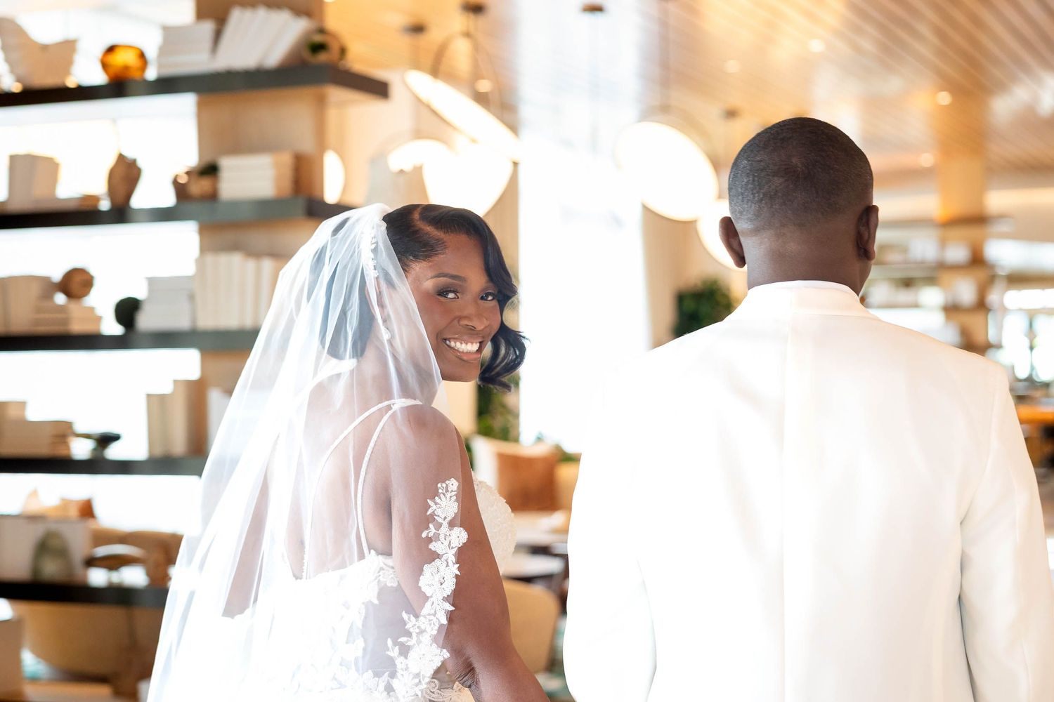 Smiling bride in lace wedding dress and veil stands with groom in bright hotel lobby.