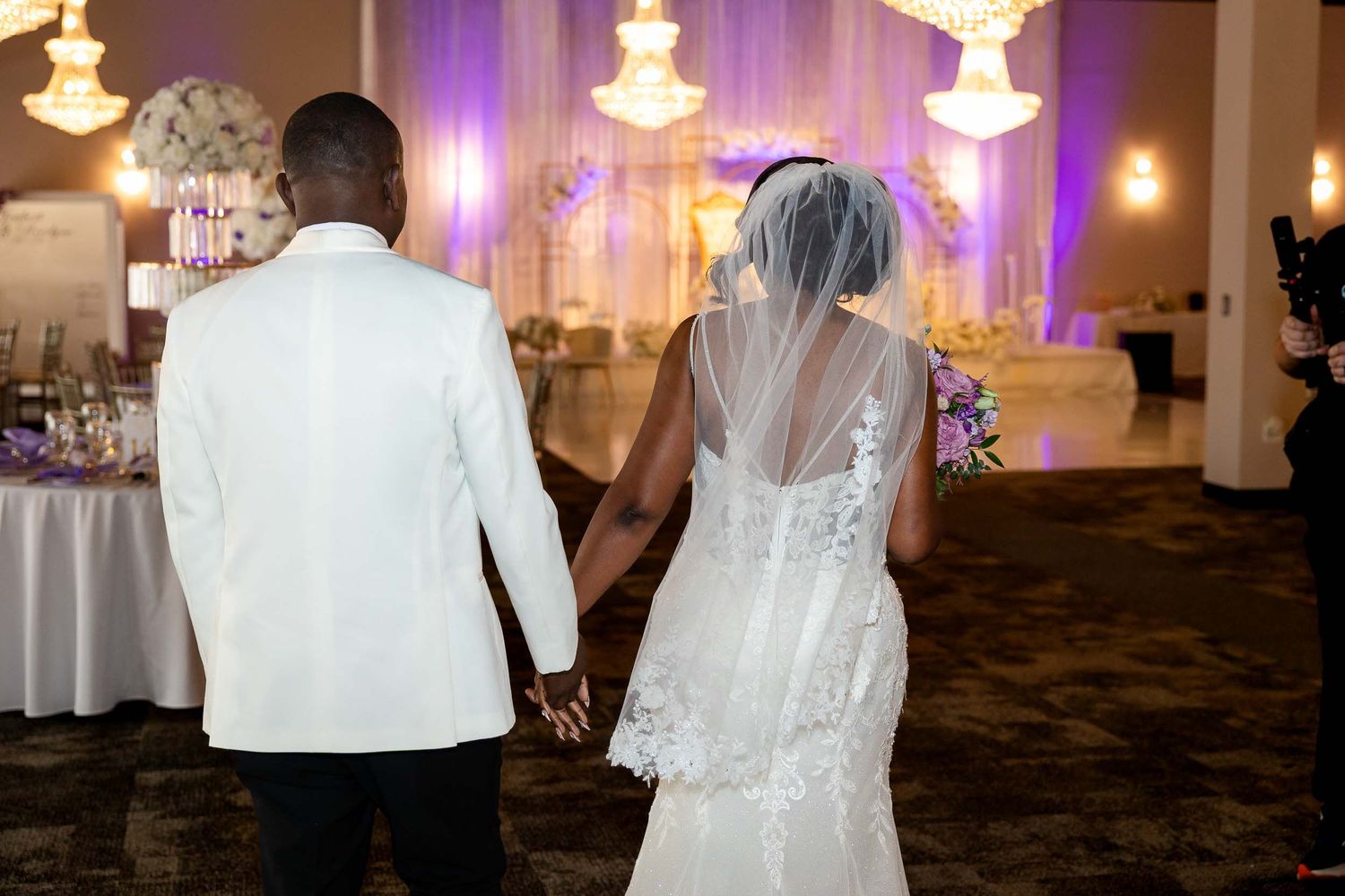Bride and groom holding hands walking into elegantly decorated wedding reception hall with purple lighting.