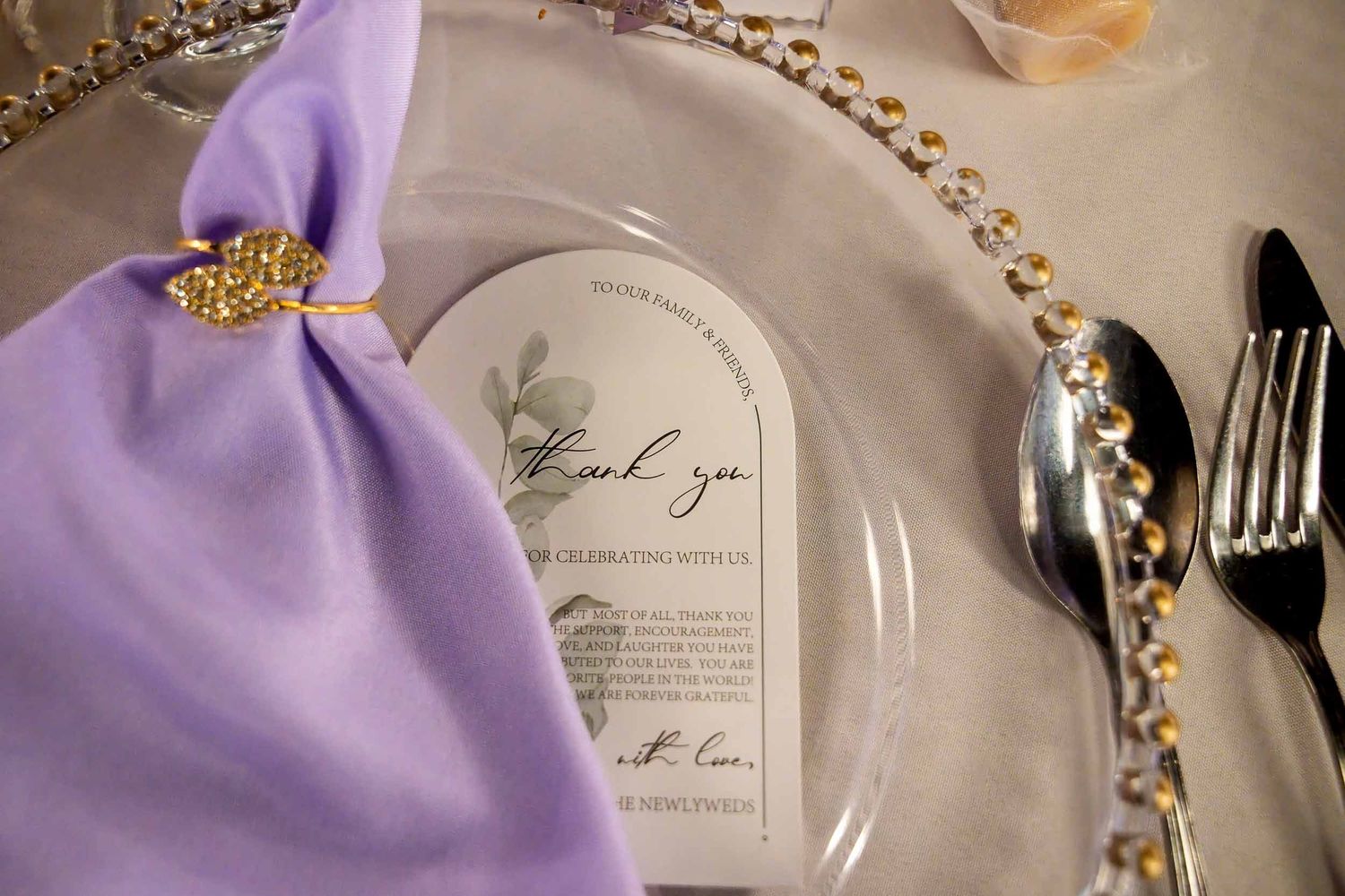 Elegant wedding place setting with lavender napkin, gold ring, silver beaded charger plate, and arch-shaped thank you card.