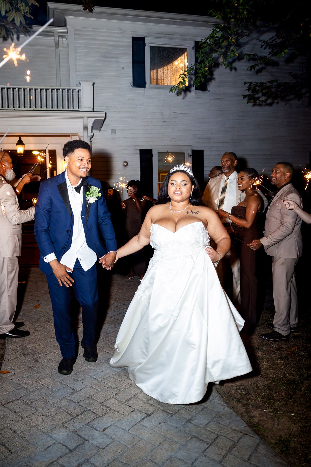 Newlywed couple exits wedding reception through sparkler sendoff, bride in white gown and groom in navy suit, night celebration.