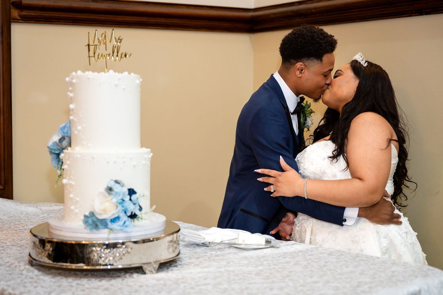 Newlywed couple sharing a kiss beside a white three-tier wedding cake with blue floral accents at their reception.