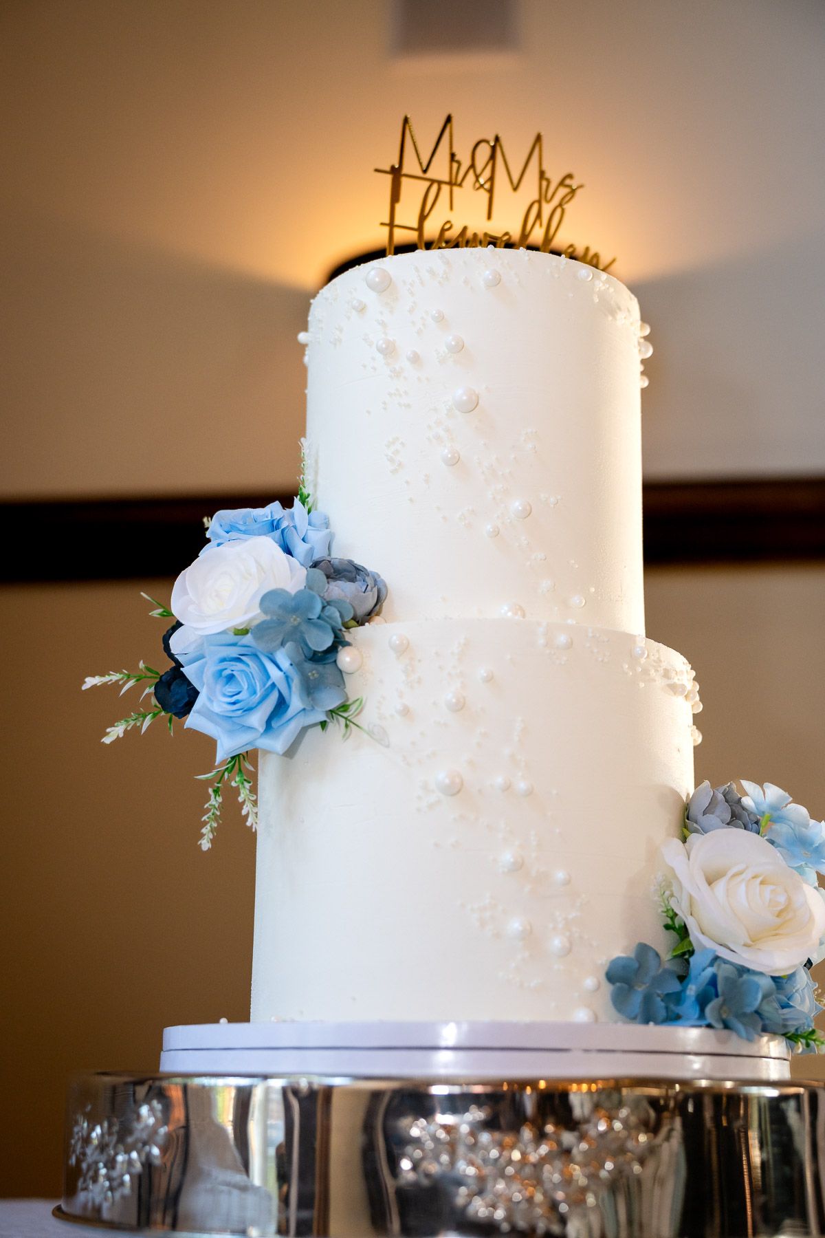 Three-tier white wedding cake with blue and white floral decorations and Mr. & Mrs. gold cake topper.
