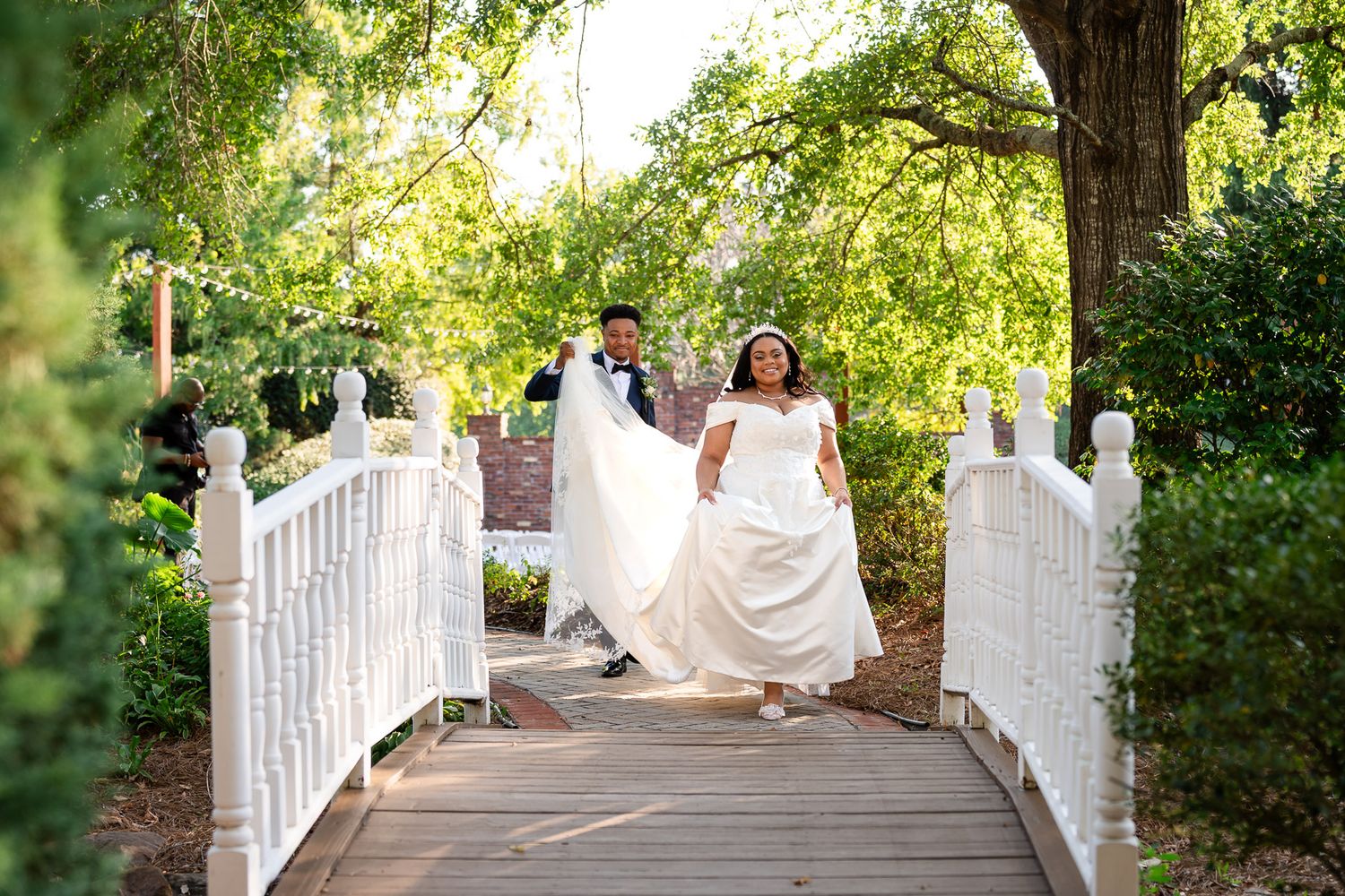 A joyful bride and groom walk hand-in-hand across a white wooden bridge surrounded by lush green trees.