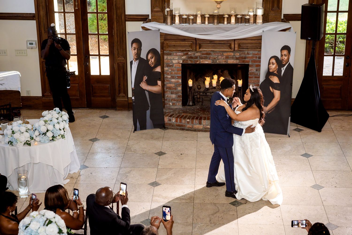 Bride and groom share their first dance at elegant wedding reception with fireplace backdrop and guests capturing the moment.