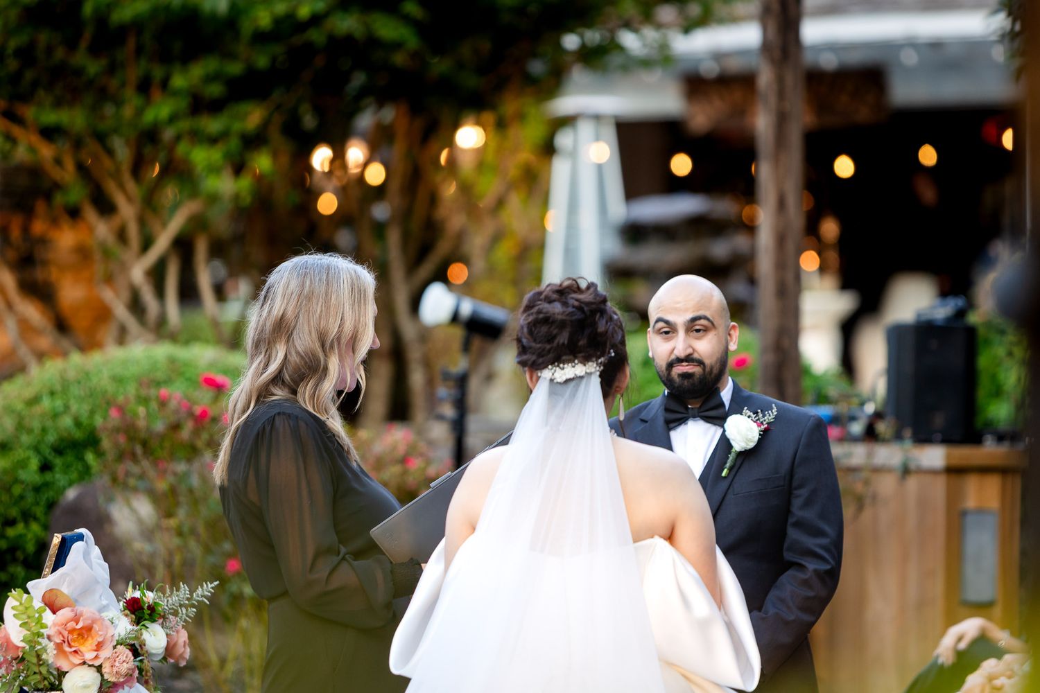 Groom in black tuxedo stands at outdoor wedding ceremony altar facing bride in white veil and officiant.