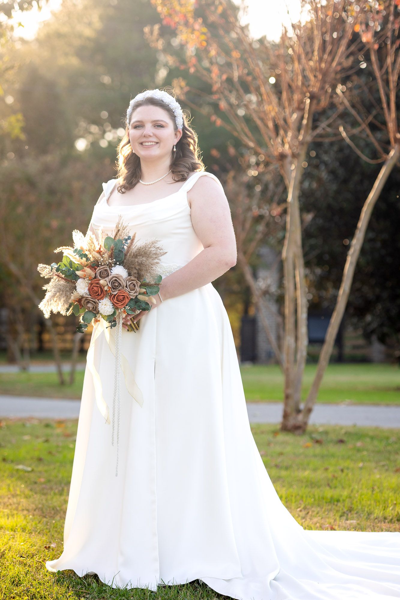 Bride in white gown holding autumn bouquet of roses and dried florals, standing outdoors in golden sunlight.