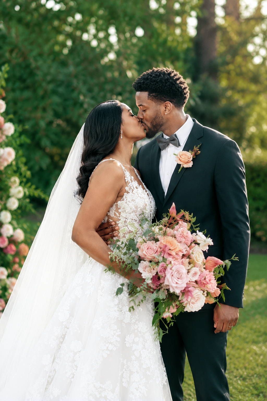 Bride and groom share a kiss outdoors, she holds a pink floral bouquet, wearing lace gown and veil, he wears a dark suit.
