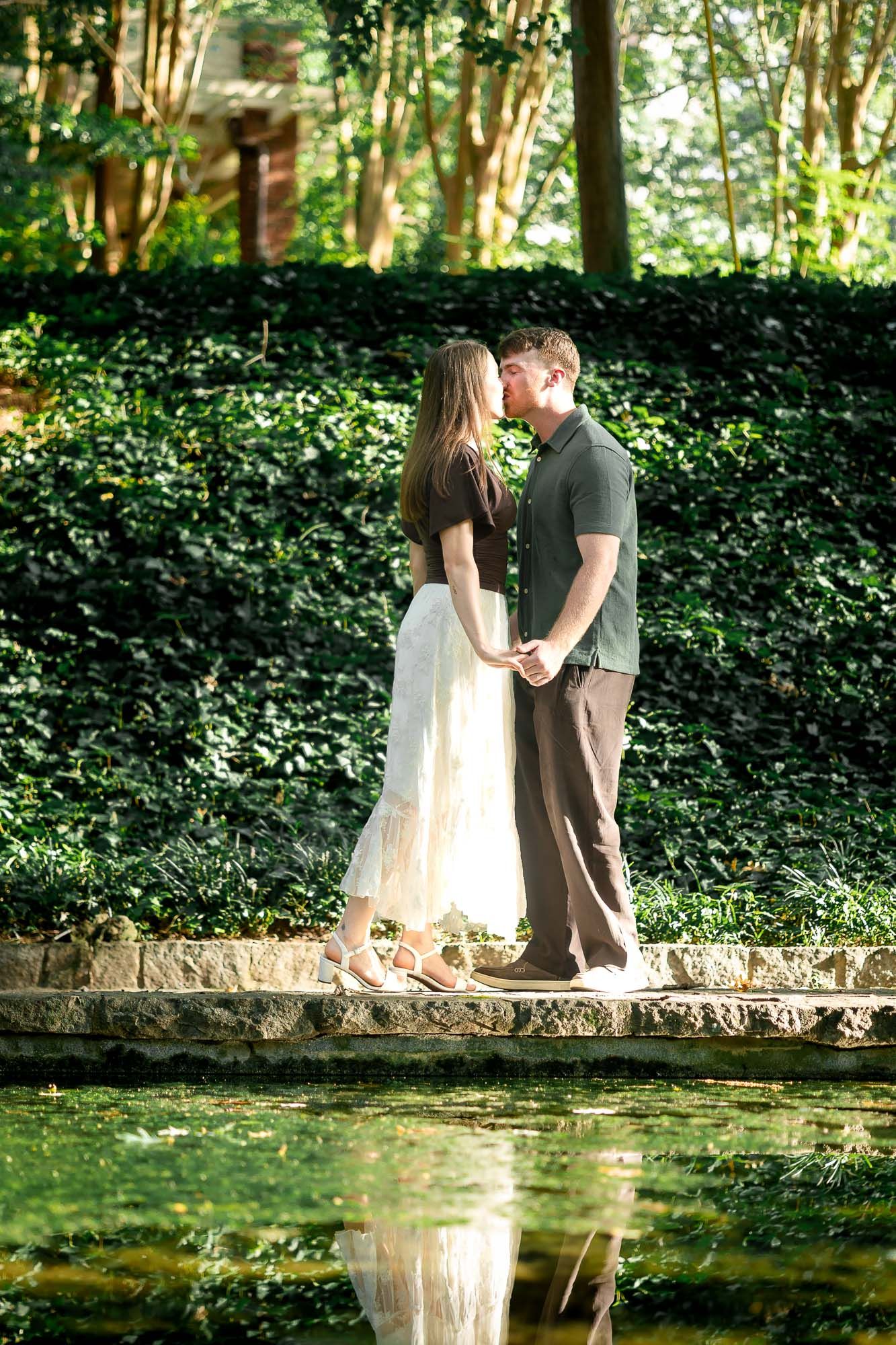 Couple kissing by a reflective pond surrounded by lush green ivy walls in a romantic garden setting.