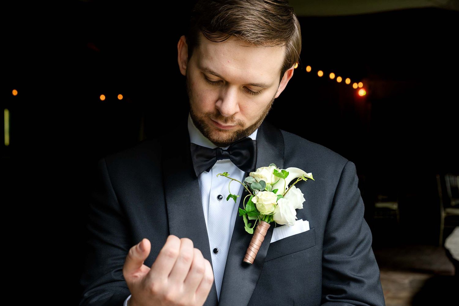 Groom in black tuxedo with bow tie adjusting boutonniere of white roses and greenery at wedding reception.