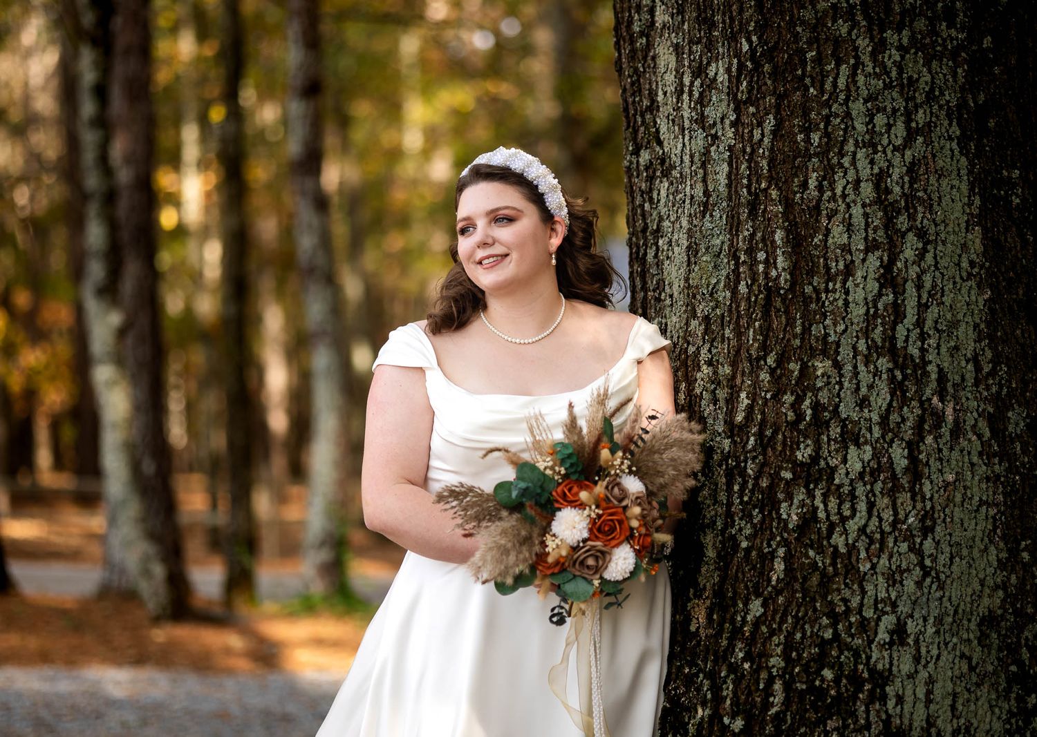 Bride in white gown holding autumn bouquet leans against tree in forest setting.