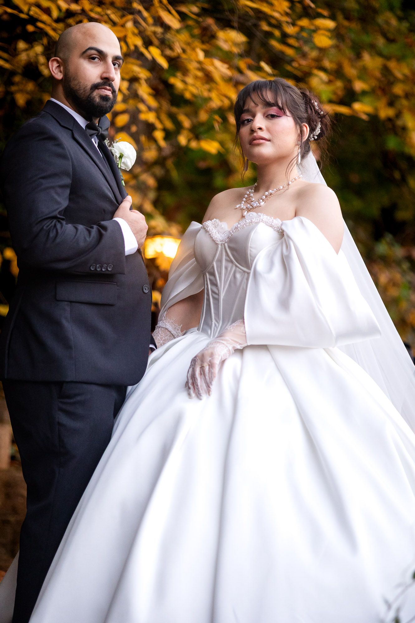 Elegant bride in white ball gown and groom in dark suit pose together outdoors among autumn foliage.
