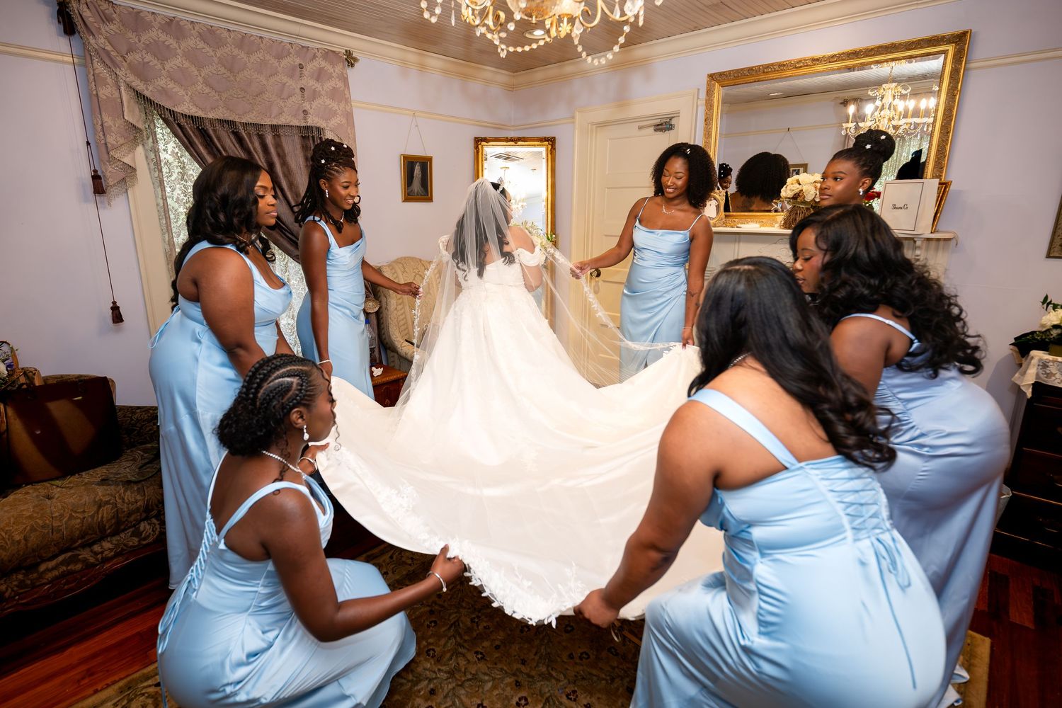 Bridesmaids in light blue dresses help a bride with her white wedding gown and veil in an elegant room.