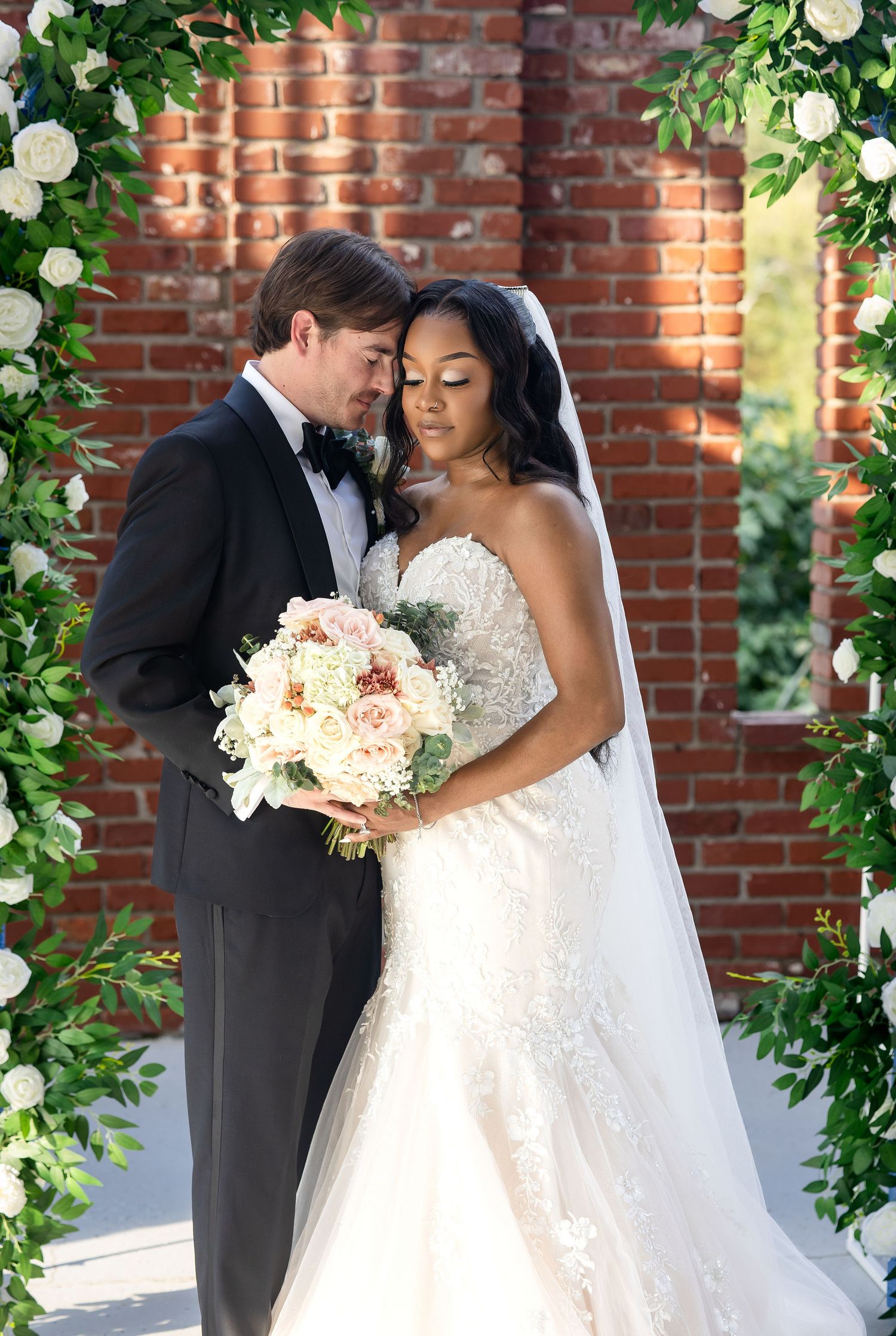 Bride and groom pose under floral arch with white roses, bride in lace gown holding bouquet, groom in black tuxedo.