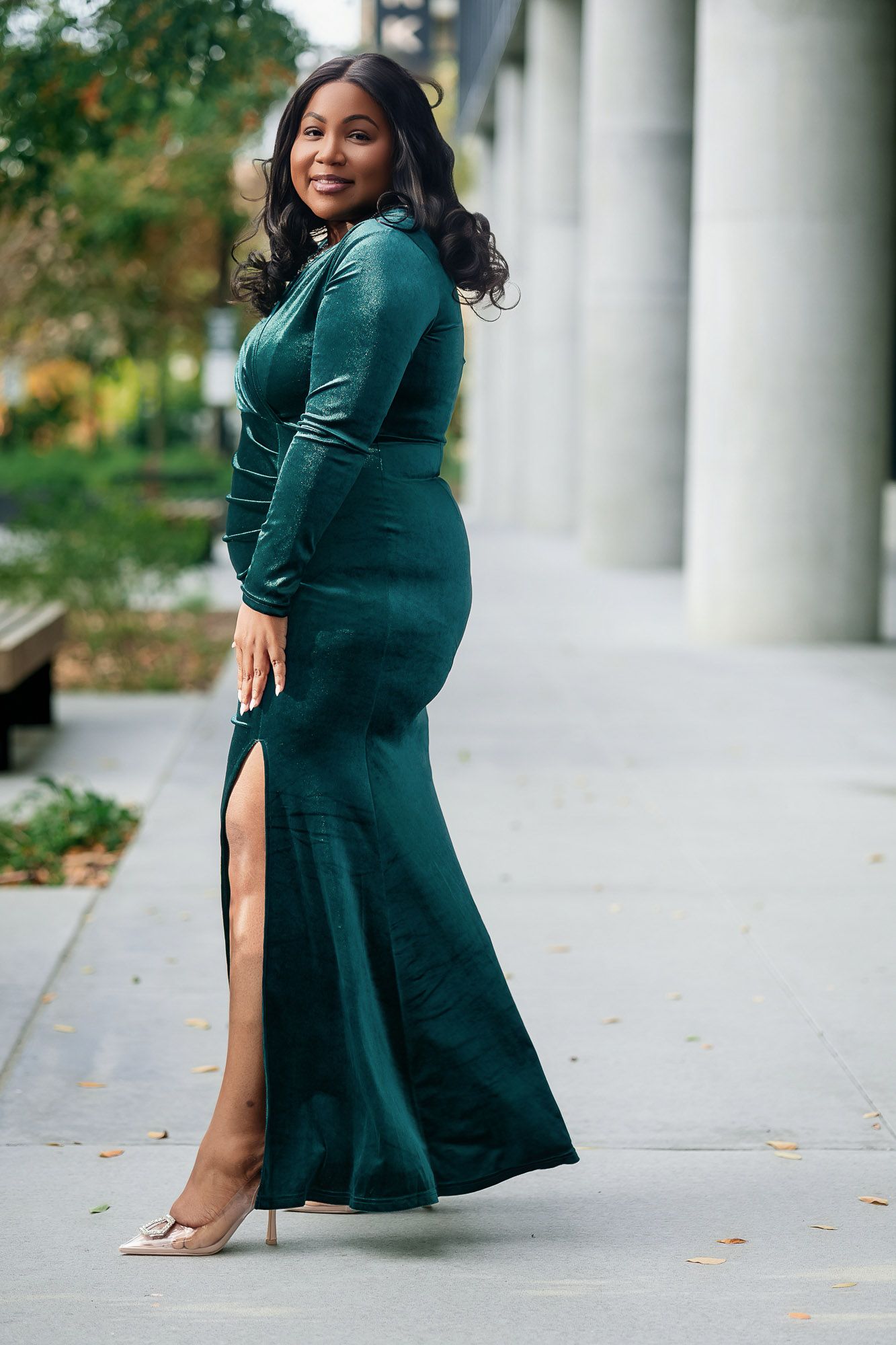 Woman in elegant emerald green velvet long-sleeve gown with thigh slit and nude heels posing outdoors.