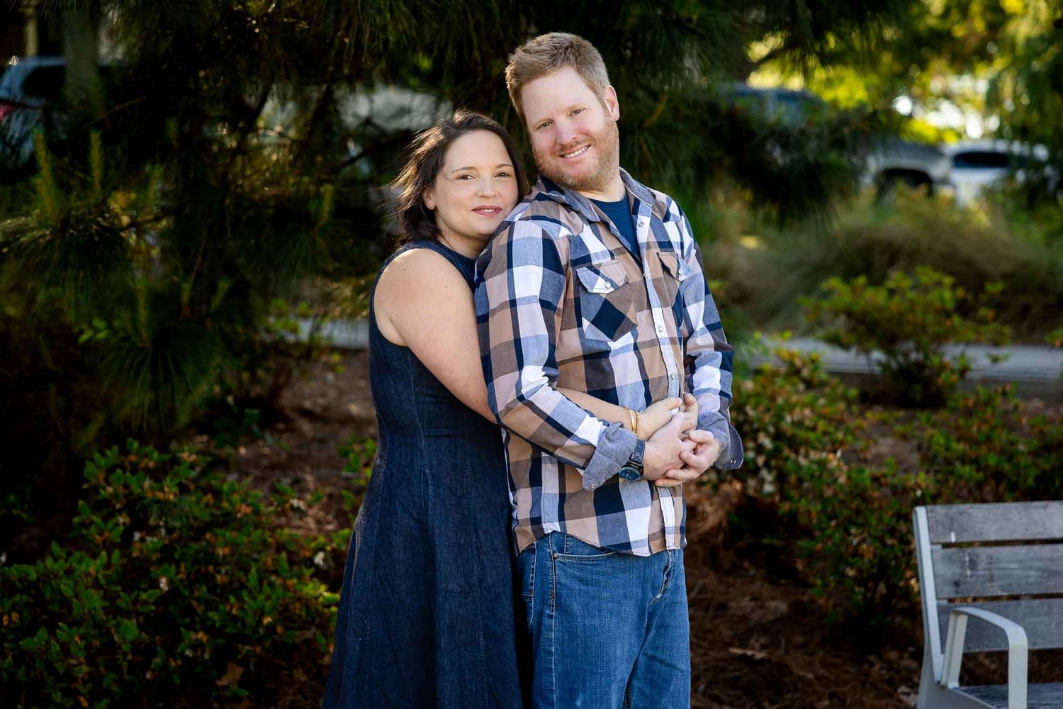Couple embracing outdoors in a lush garden setting, woman in navy dress and man in plaid shirt smiling warmly.