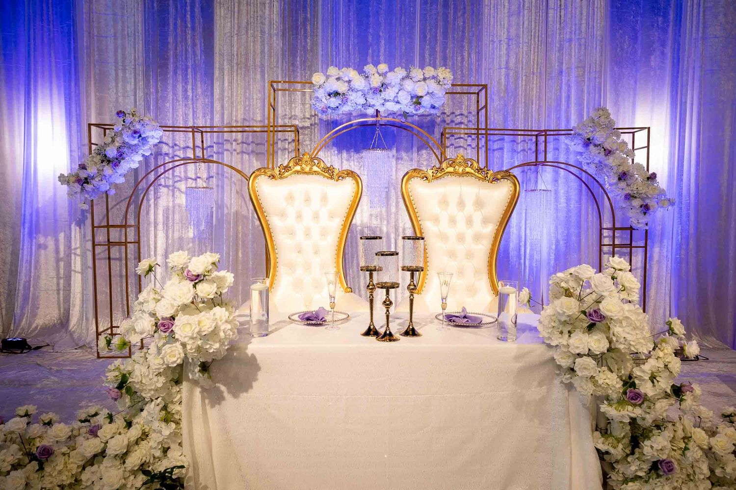 Elegant wedding sweetheart table with gold throne chairs, white floral arrangements, and purple-lit backdrop decor.