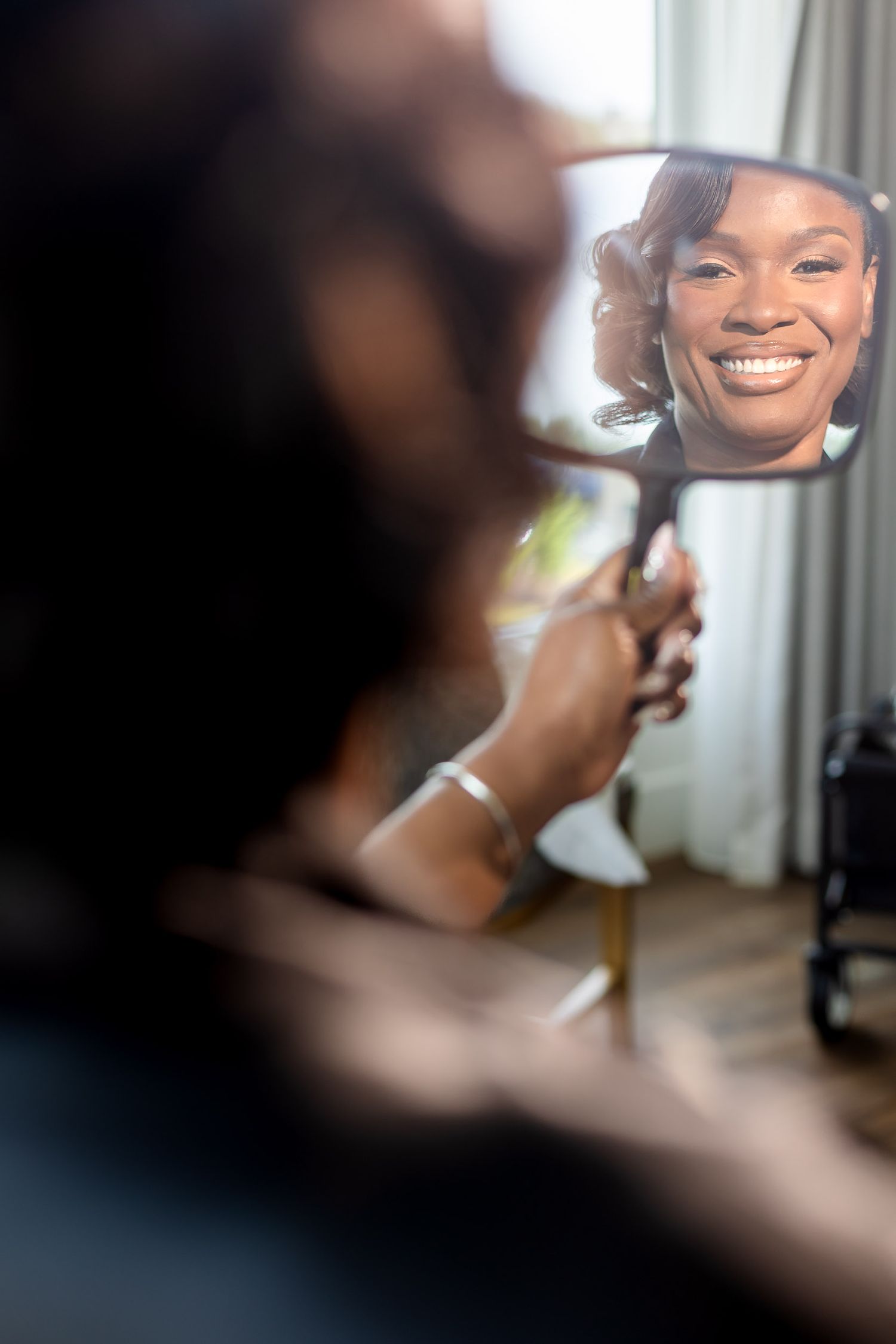 A smiling Black woman views her reflection in a handheld mirror while getting ready, showcasing elegant hair and makeup.