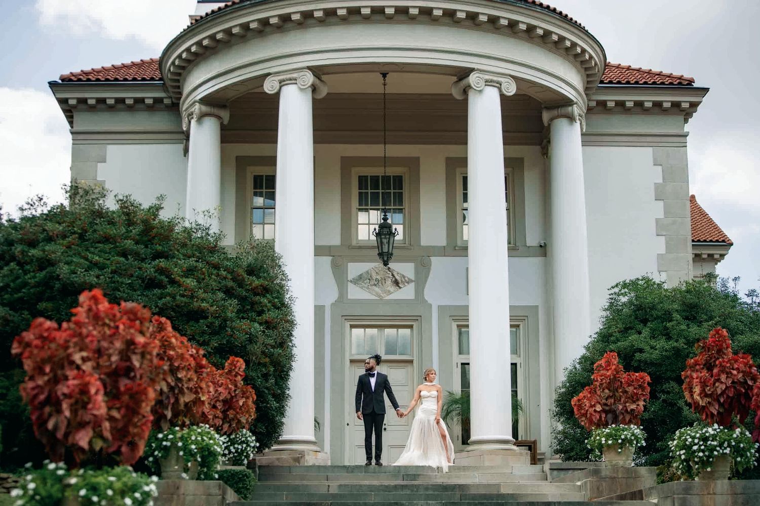 Bride and groom walk down steps of elegant white columned mansion surrounded by lush red foliage on wedding day.
