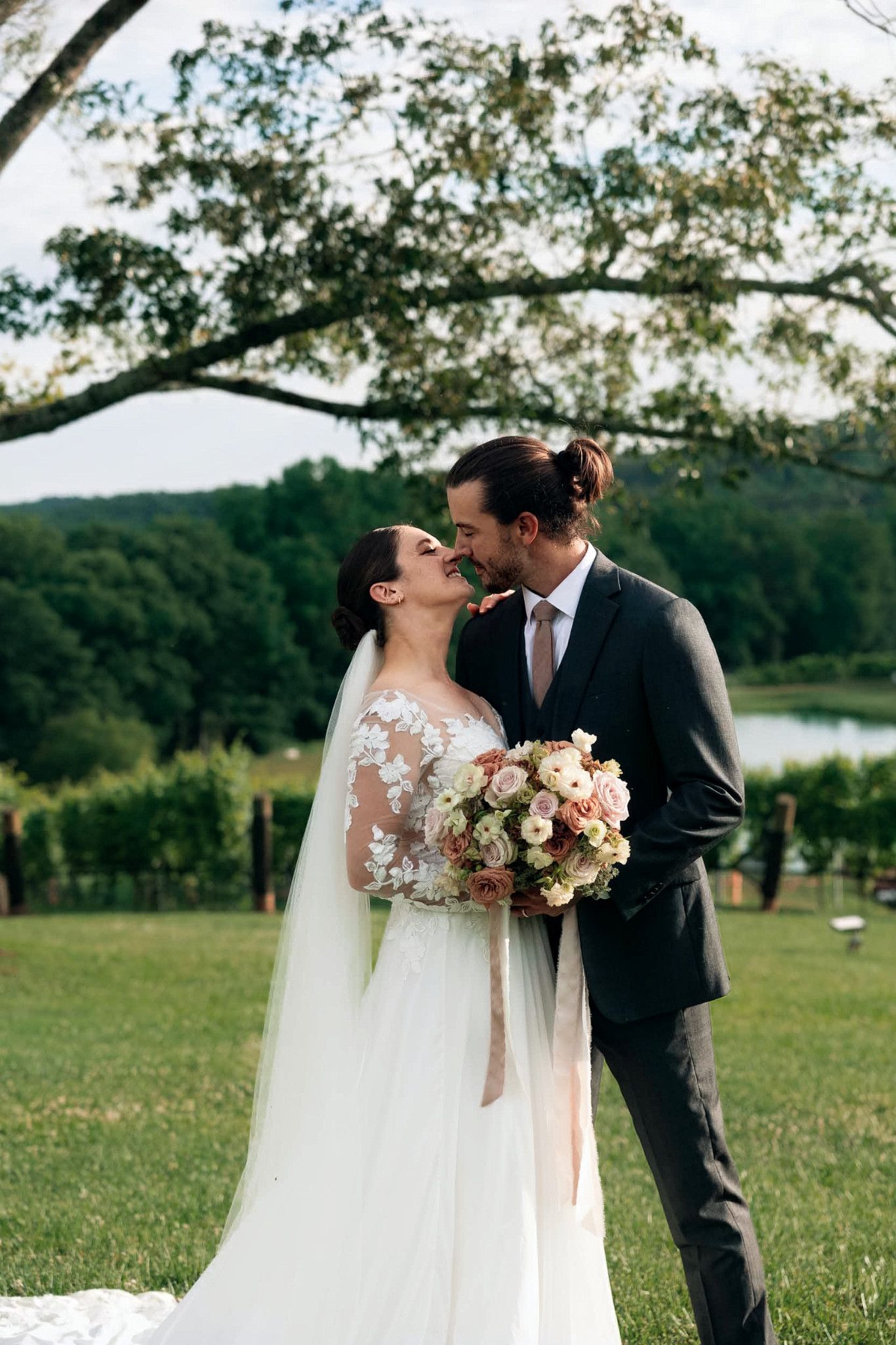 Bride and groom share a romantic kiss outdoors at a vineyard wedding, bride holding a pastel bouquet in a lace gown.