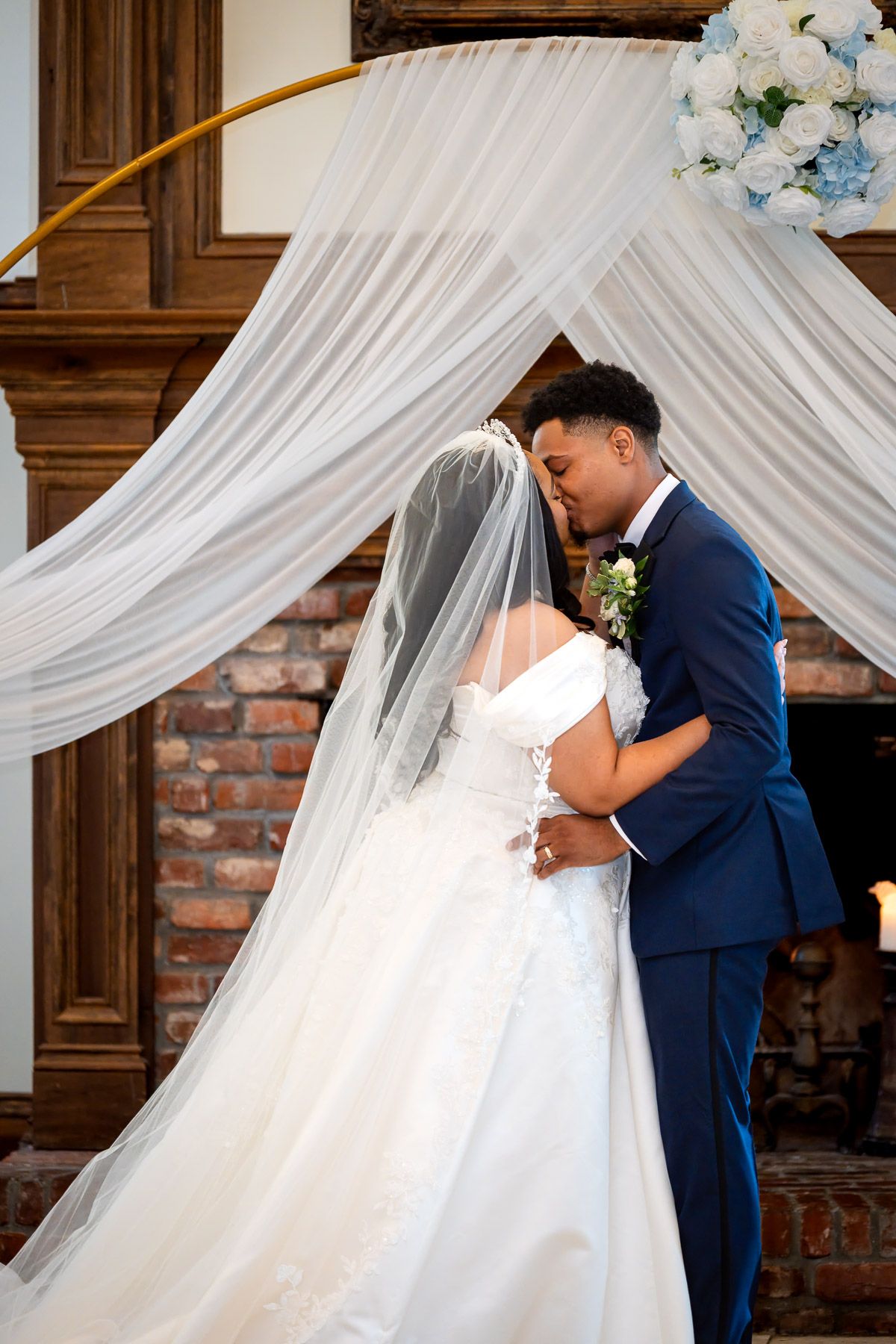 Bride and groom share first kiss under white draped arch with blue flowers at elegant indoor wedding ceremony.
