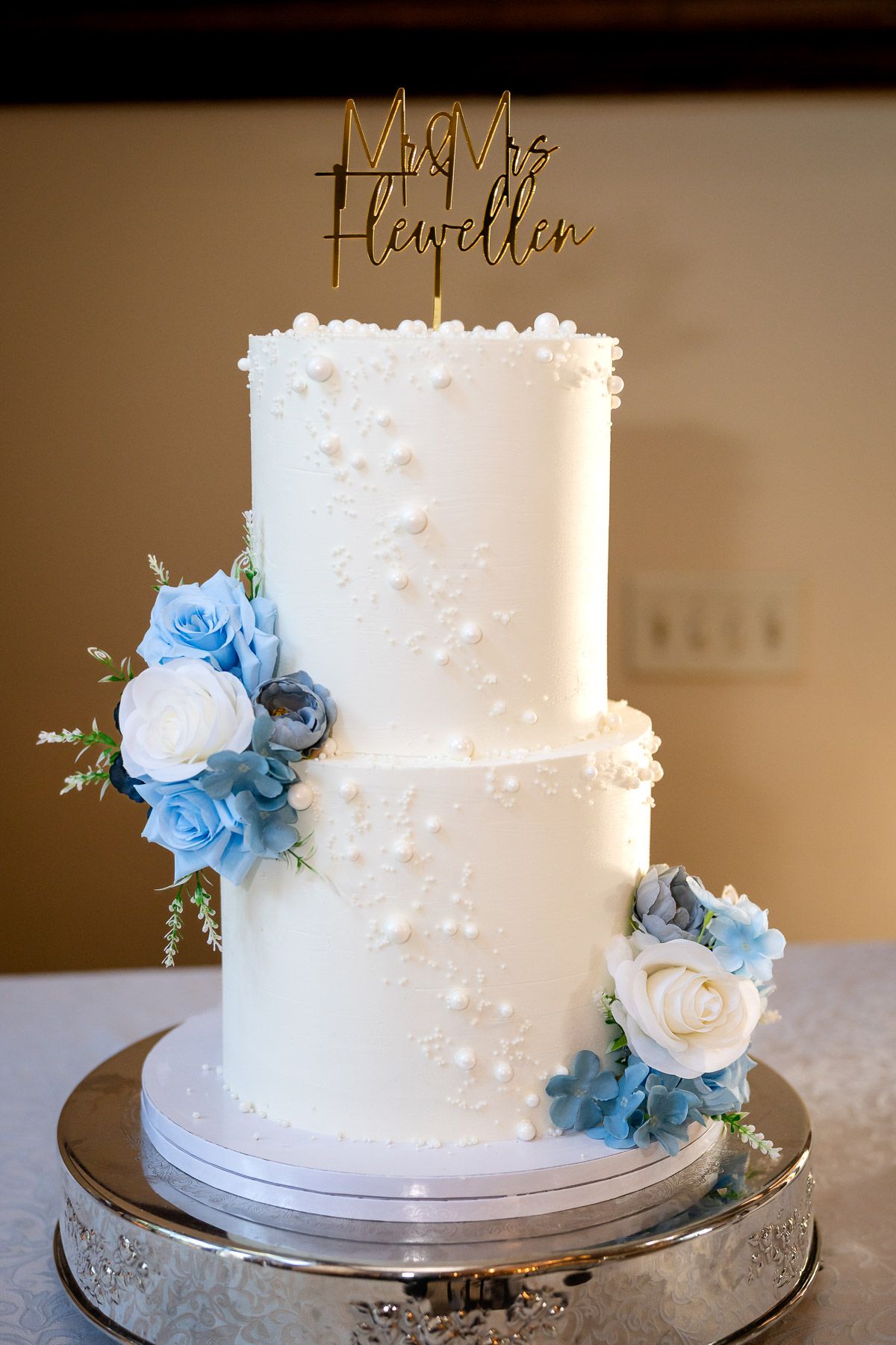 Two-tier white wedding cake with blue and white floral accents and gold Mr. & Mrs. Flewellen cake topper on silver stand.