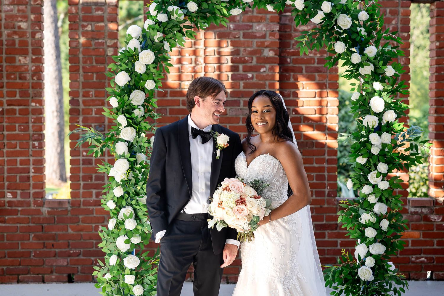 Bride and groom smiling under a floral arch with white roses and greenery against a brick wall backdrop.