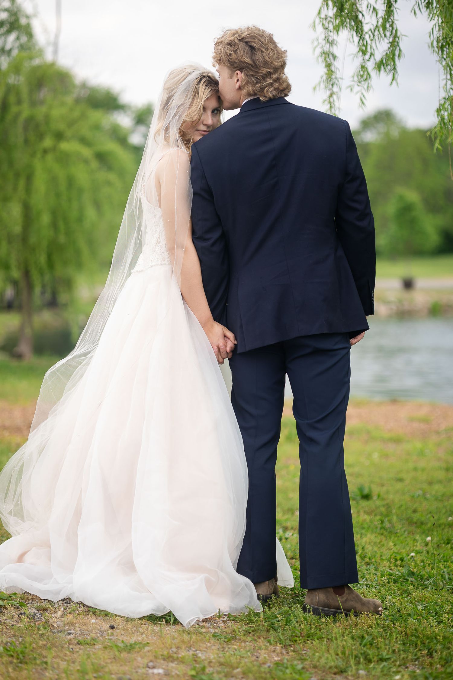 A romantic wedding photo showing a couple standing by a lake with weeping willow trees in the background.