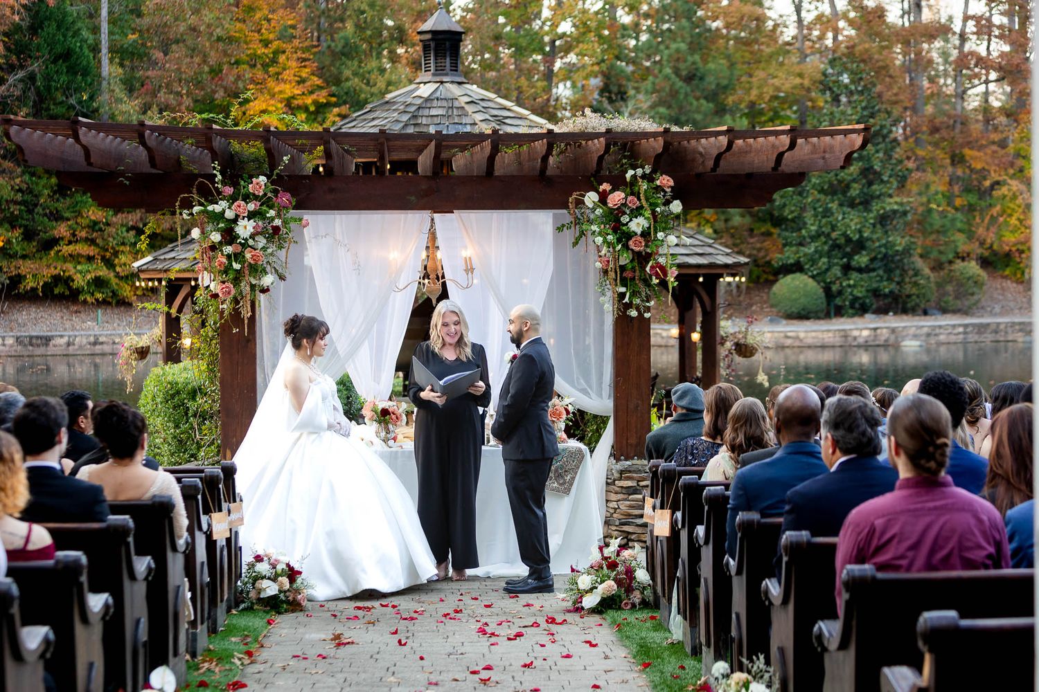 Outdoor wedding ceremony at a gazebo with fall foliage, bride and groom exchanging vows before seated guests.
