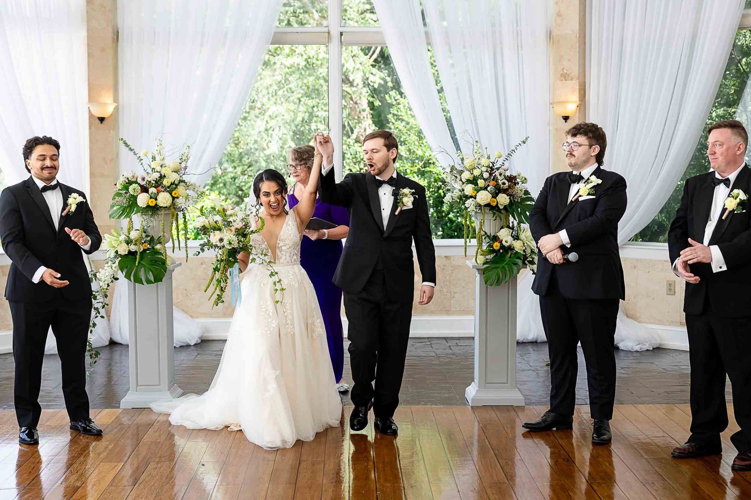 Joyful newlyweds raise hands at elegant indoor wedding ceremony with groomsmen and floral arrangements.