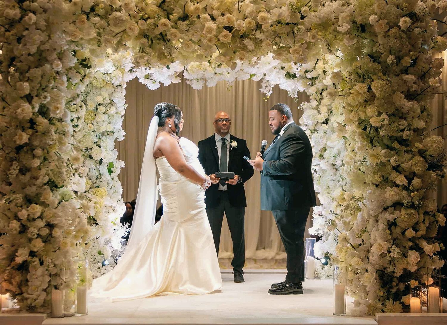 Bride and groom exchange vows under an elegant floral arch adorned with white flowers at a luxurious wedding ceremony.
