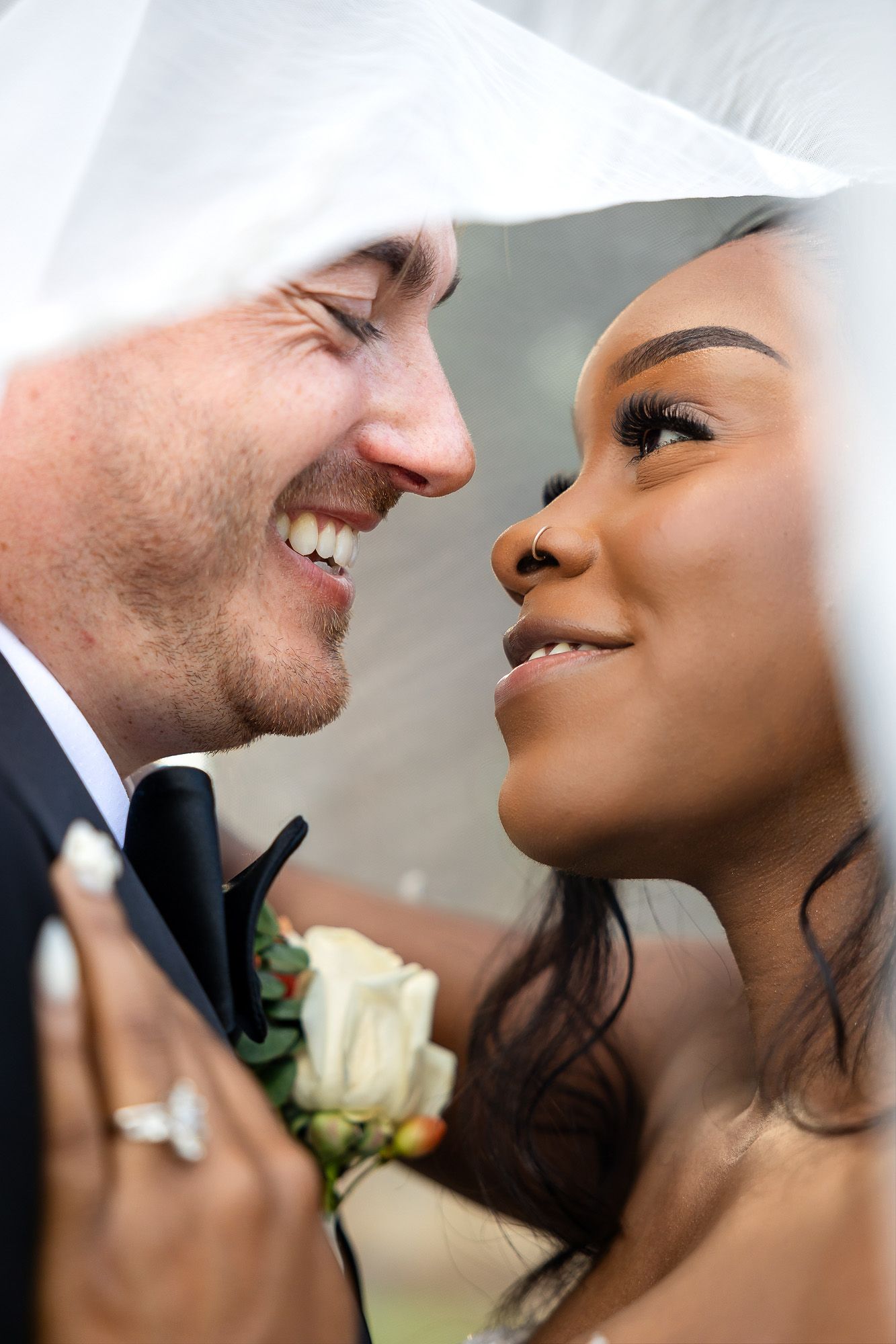 Interracial newlywed couple sharing a romantic moment under bridal veil, groom in tuxedo, bride holding white roses.