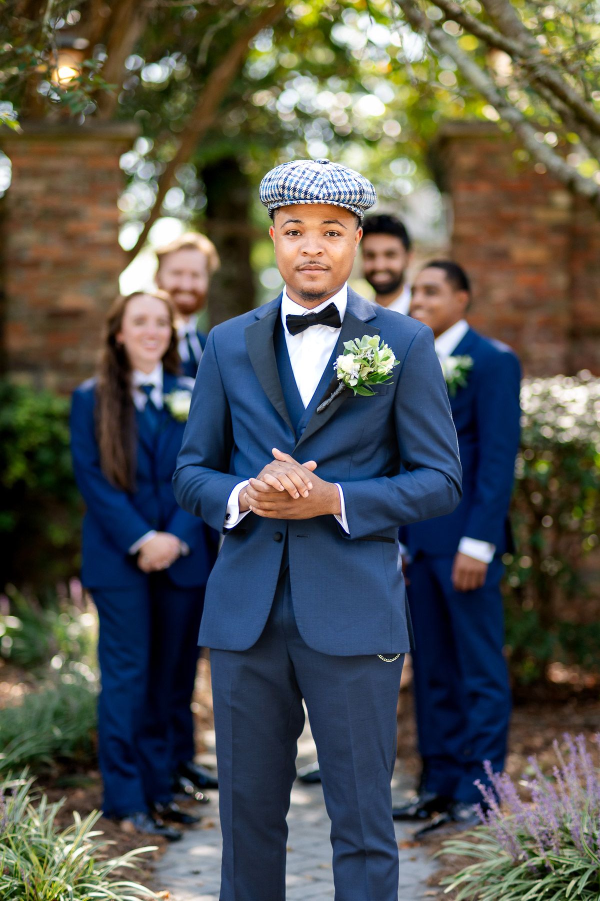 Groom in navy suit and plaid flat cap with boutonniere stands before wedding party in matching blue suits outdoors.