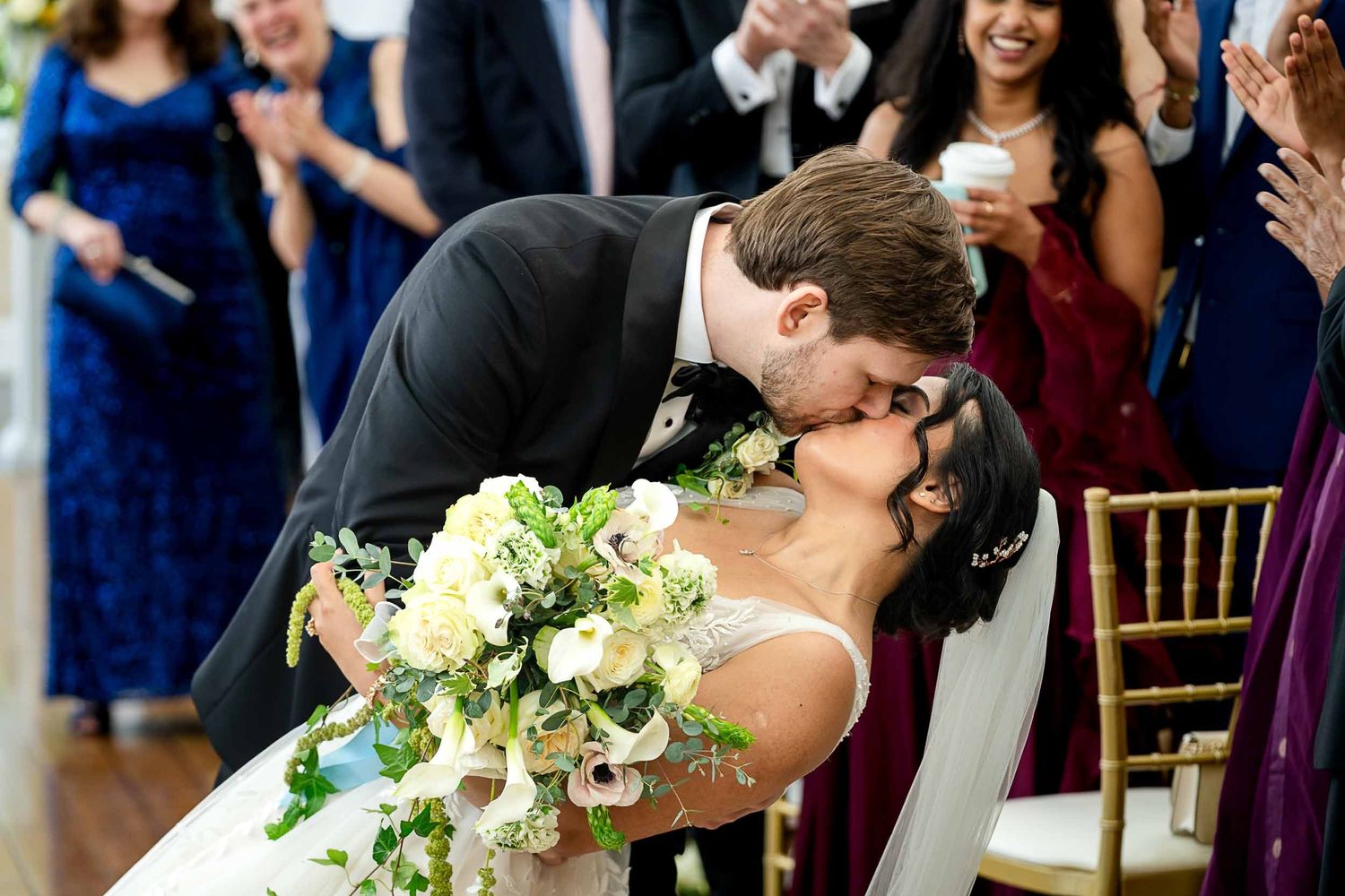 Bride and groom share a romantic dip kiss at wedding ceremony while guests applaud in elegant venue.