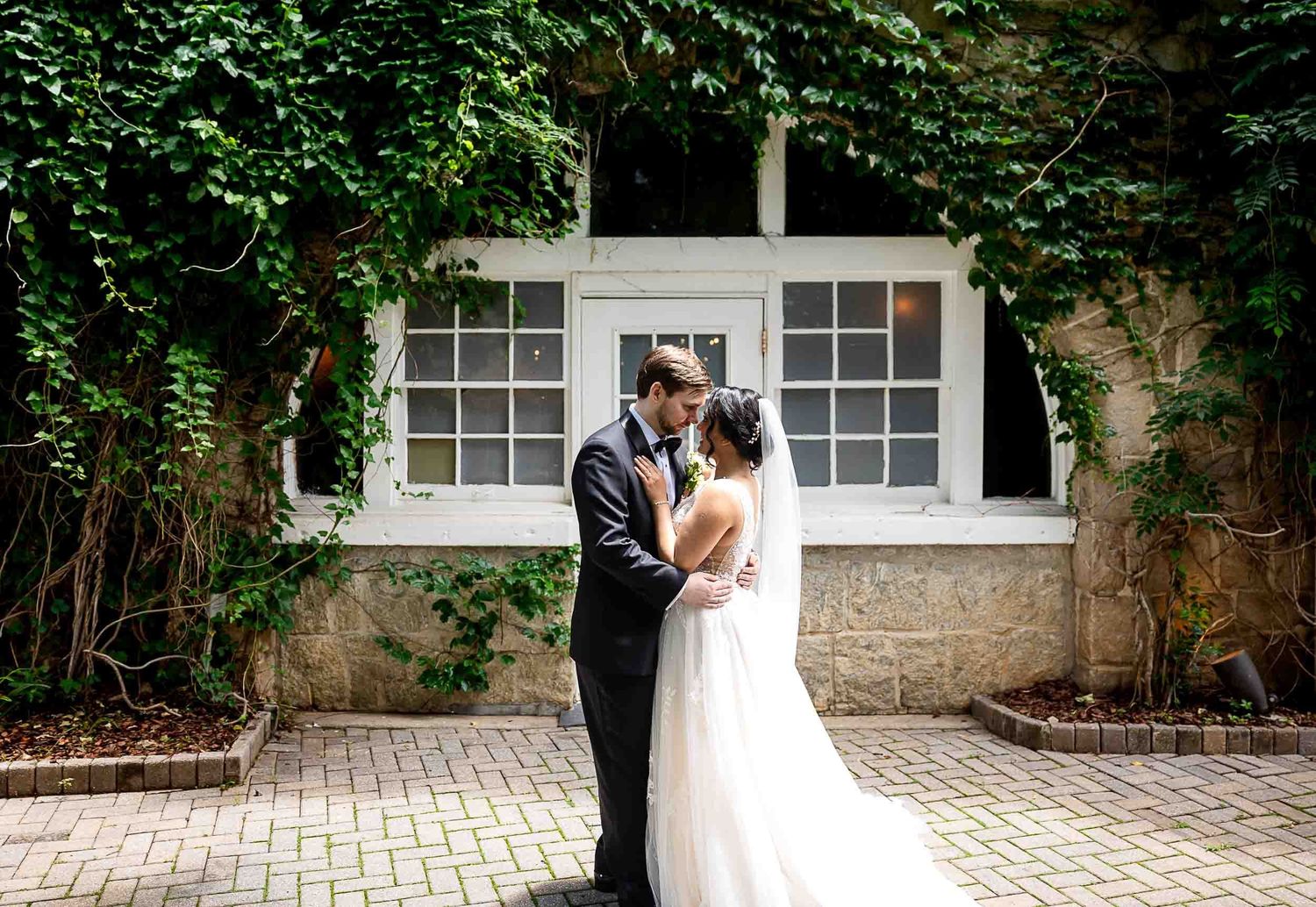 Bride and groom share a romantic embrace outdoors, framed by lush ivy-covered walls and a charming white window.