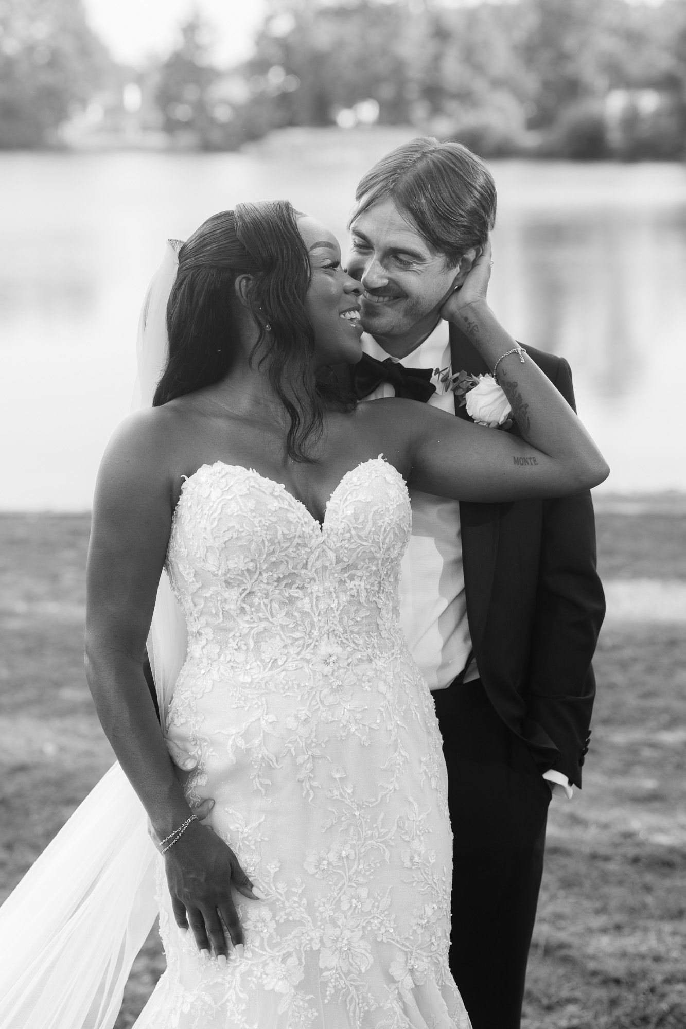 Black and white photo of an interracial couple in wedding attire sharing a romantic moment near a lake.