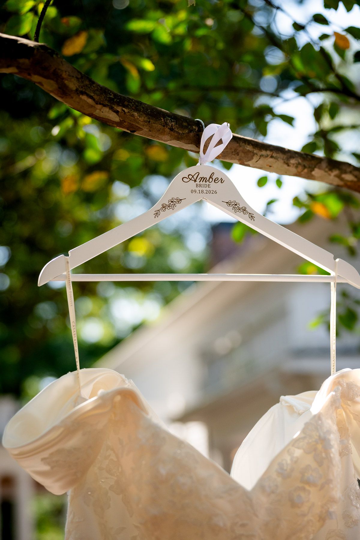 Personalized white wedding dress hanger with bride's name hanging on a tree branch outdoors.