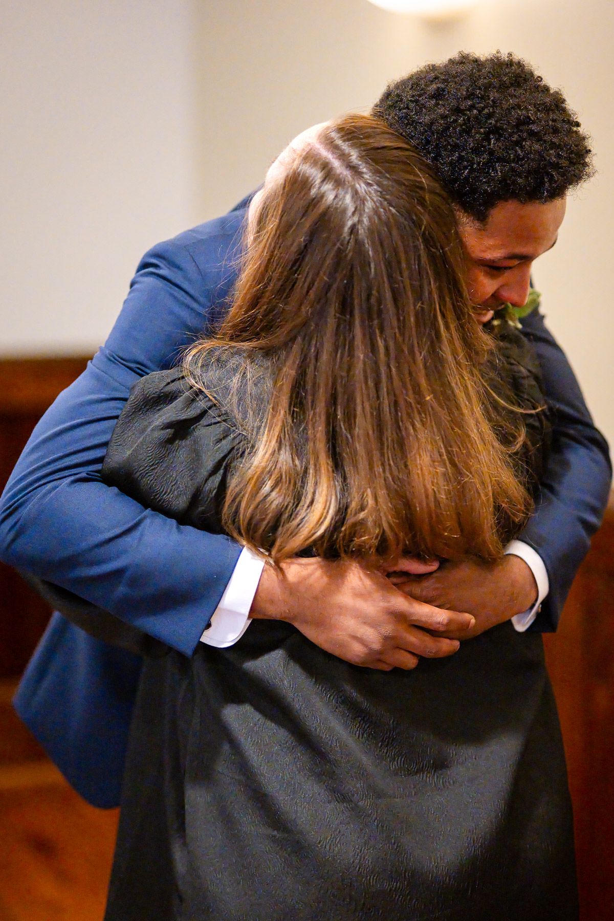 A man in a blue suit embraces a woman in a black dress, sharing a heartfelt hug indoors.