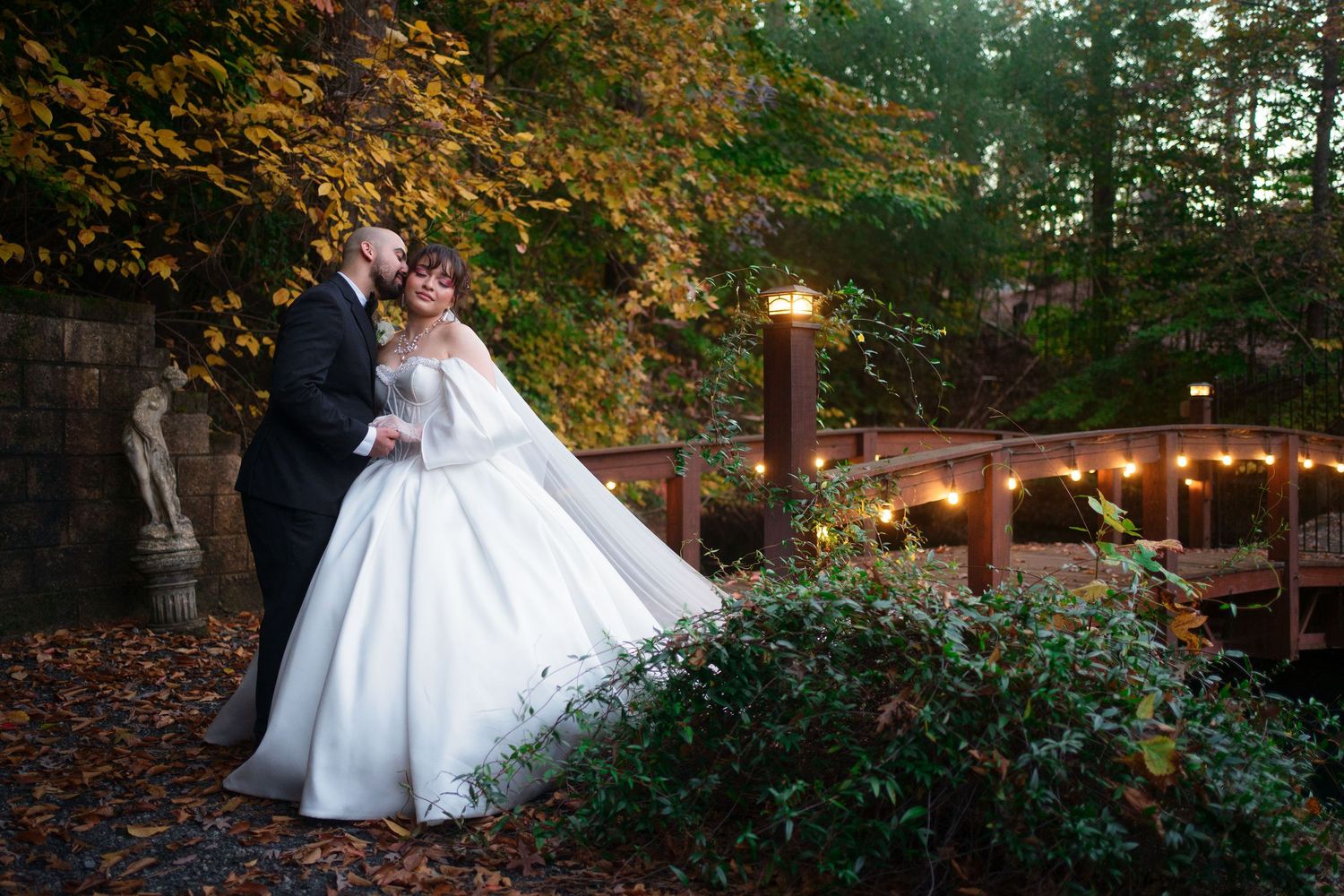 Bride and groom share romantic kiss in autumn garden with string lights and colorful foliage at outdoor wedding venue.