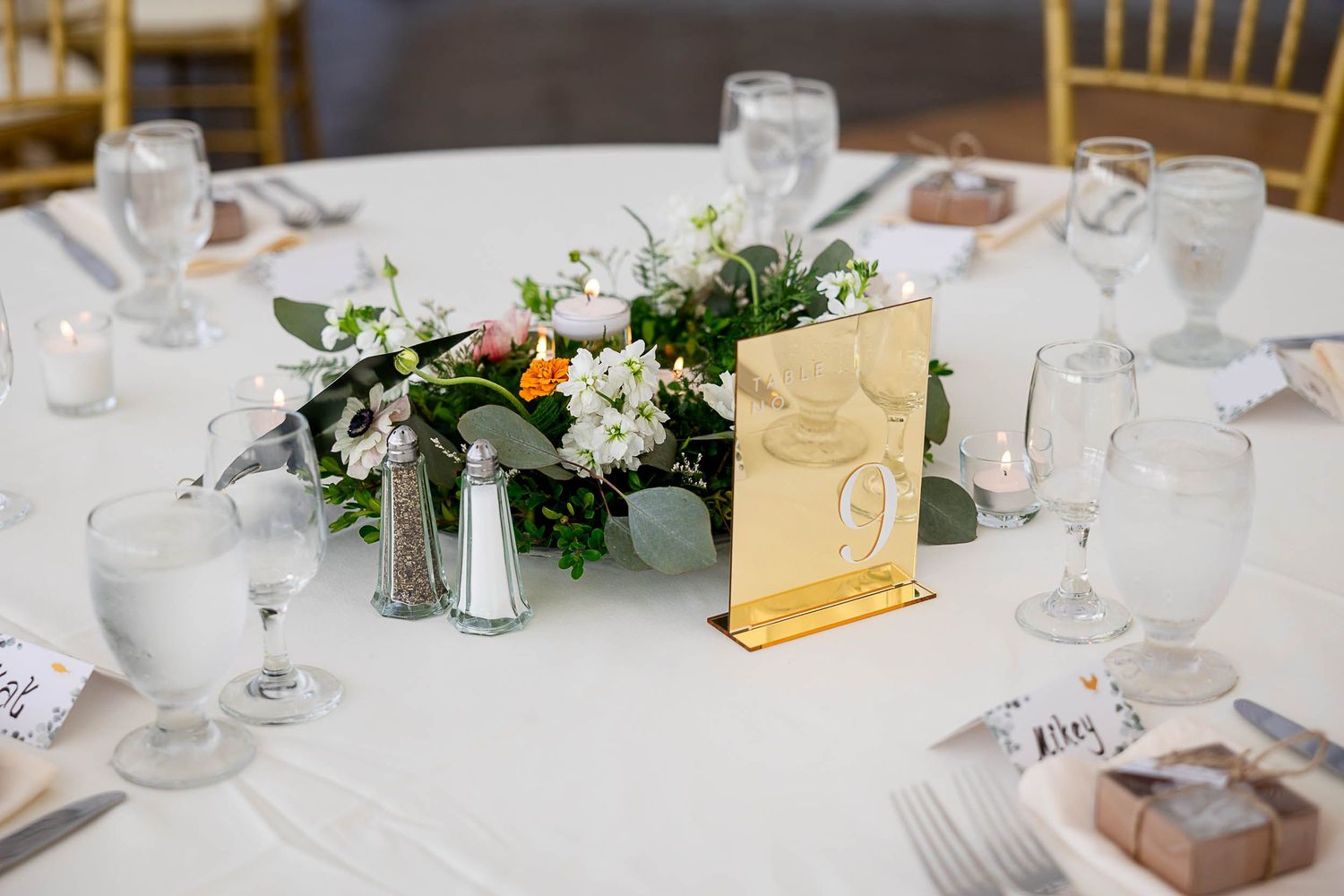 Elegant wedding reception table with floral centerpiece, gold table number 9, white linens, and glassware.