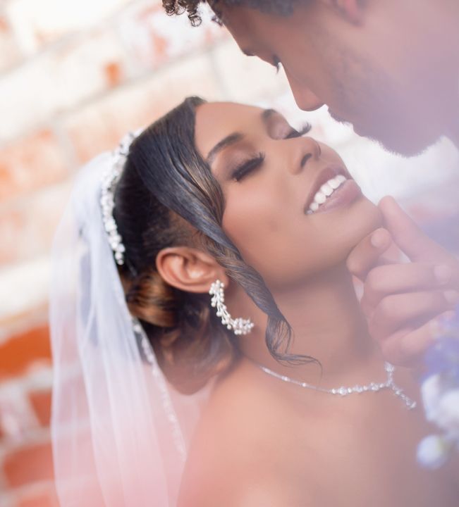 Bride in white veil and pearl accessories smiles as groom gently holds her chin during romantic wedding moment.
