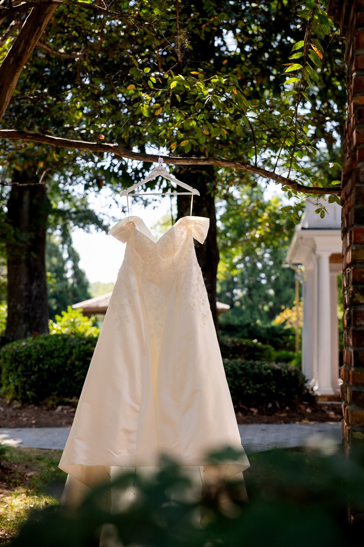 Elegant off-shoulder white wedding dress hanging on a hanger outdoors among lush green trees near a classic white building.