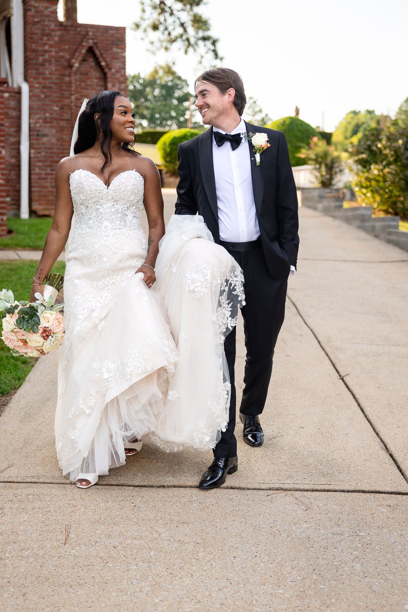 A bride and groom walk together outside a church, she in a white lace gown holding flowers, he in a black tuxedo.