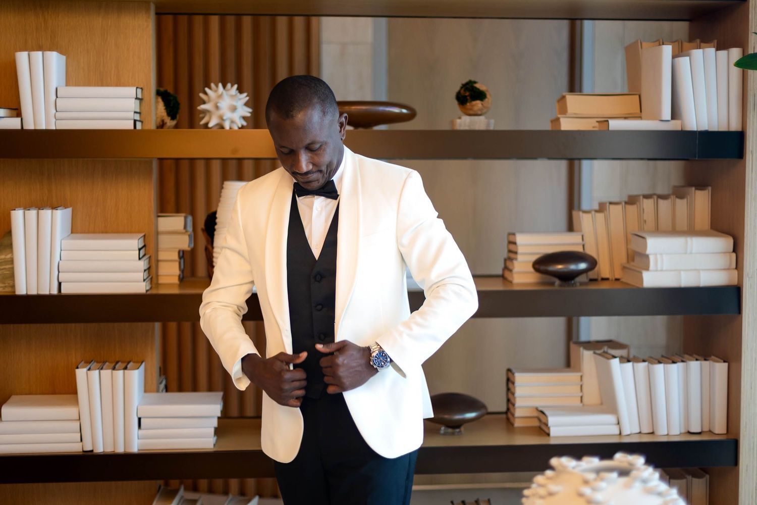 Elegant man in white tuxedo jacket and bow tie standing before a styled bookshelf with white books.