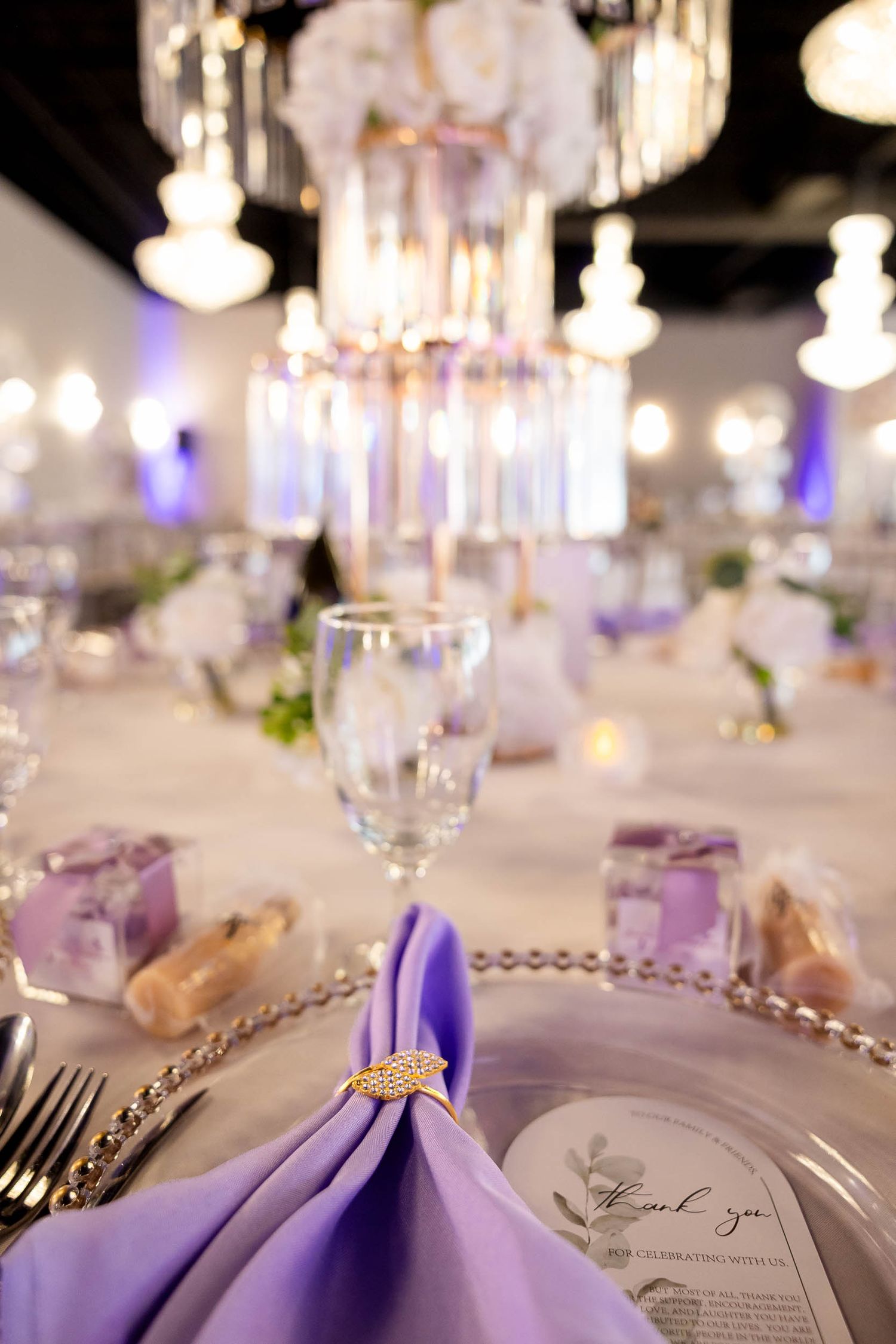 Elegant wedding reception table with lavender napkin, crystal charger plate, and glowing chandeliers in a purple-lit venue.