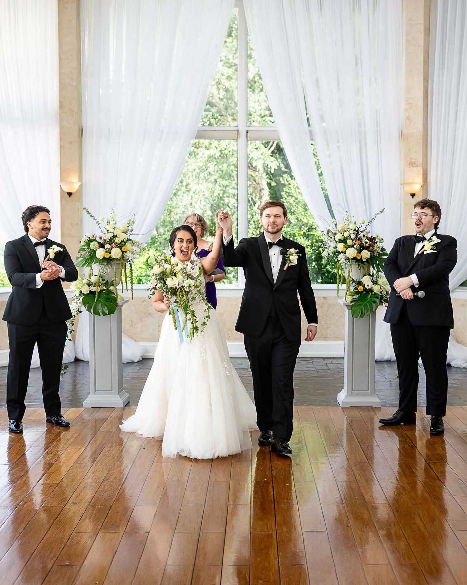 Joyful newlyweds celebrate at wedding altar with groomsmen, white floral arrangements, and sheer curtains in elegant venue.