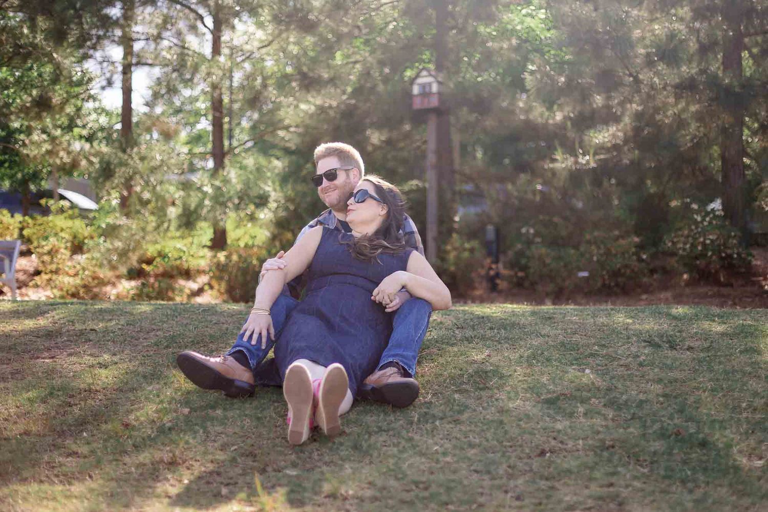 Couple sitting together on green grass in a sunny park, surrounded by trees, both wearing sunglasses and casual clothes.
