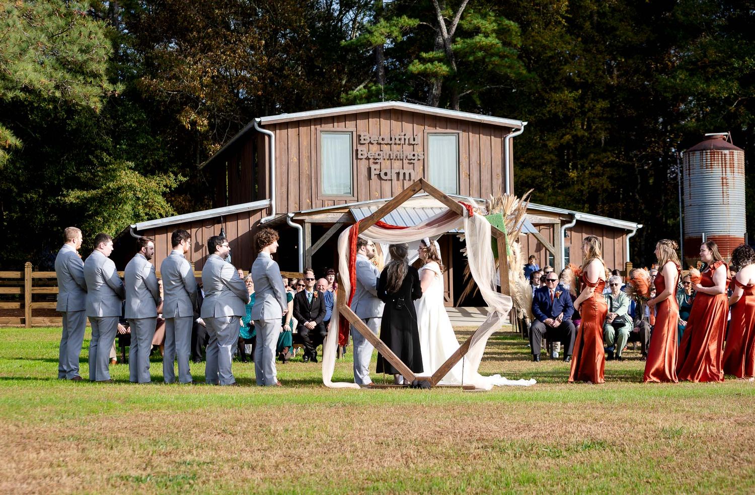 Outdoor wedding ceremony on green lawn with bridal party in blue suits and red dresses before rustic barn venue.