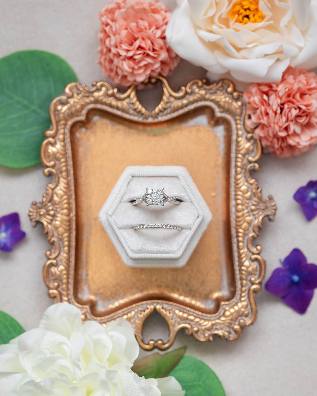 Diamond engagement ring and wedding band displayed in a hexagonal velvet ring box on a gold ornate tray with flowers.