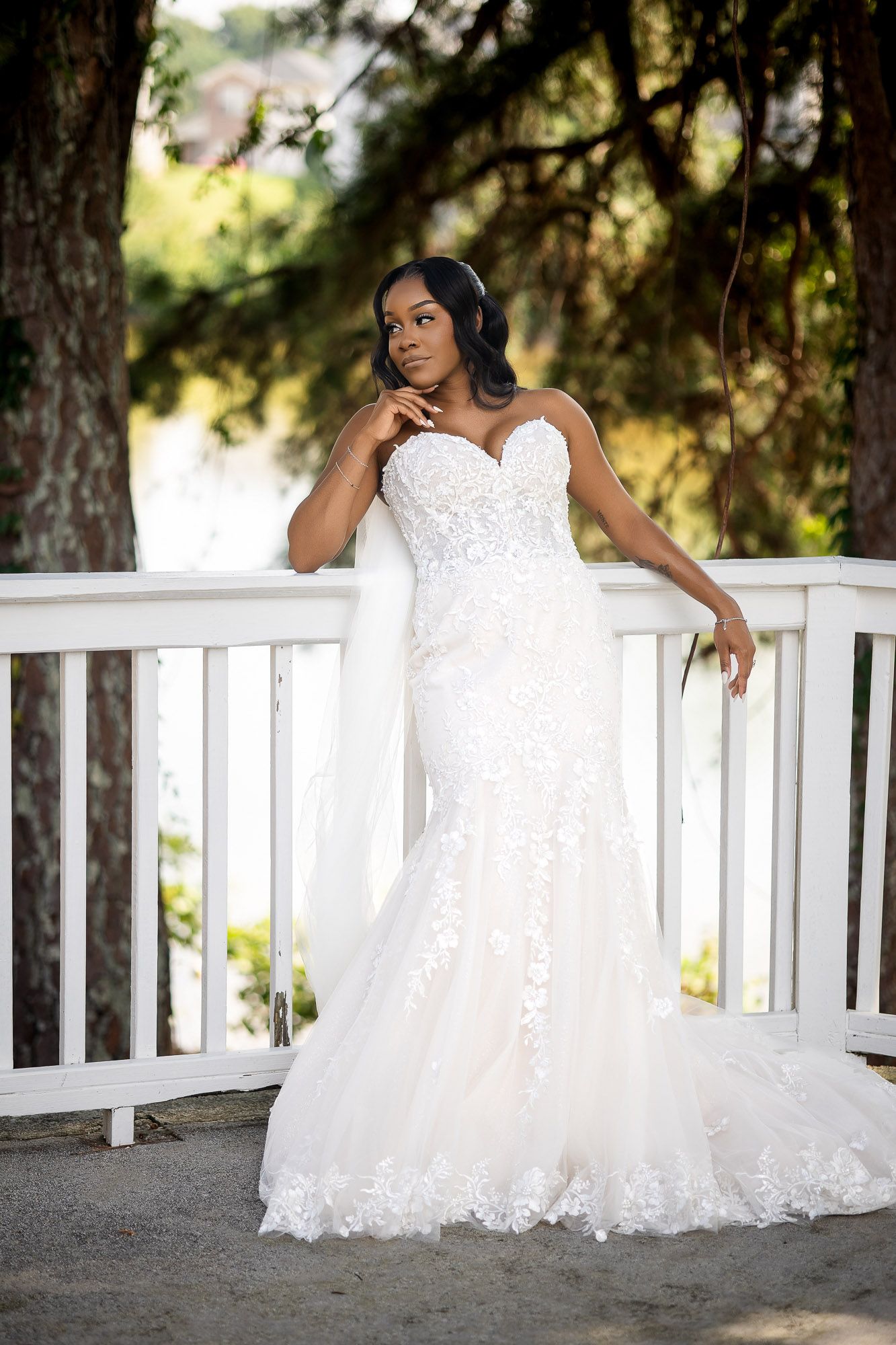 Bride in strapless lace mermaid wedding gown with veil poses gracefully on white porch railing outdoors.